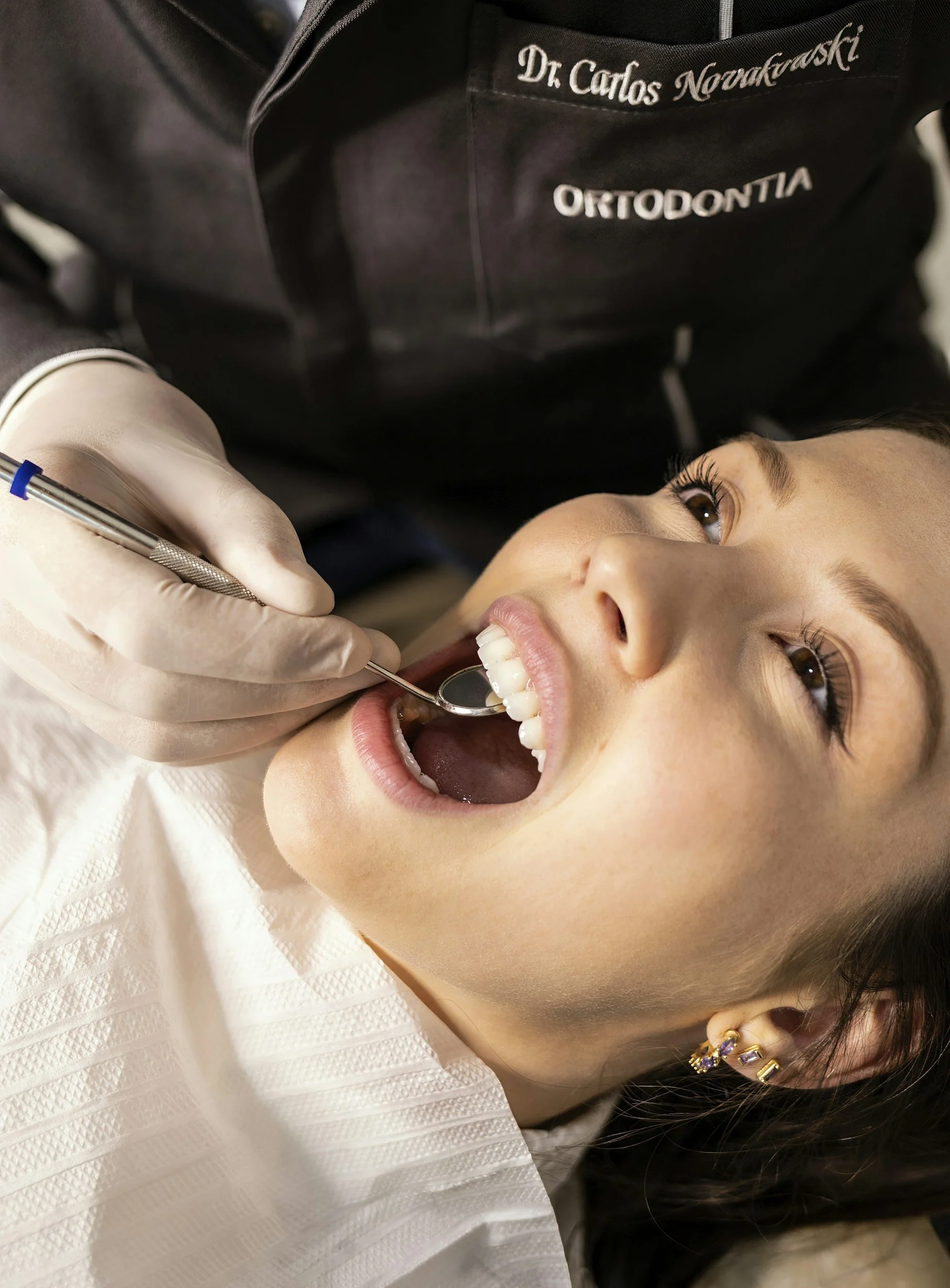 A dentist wearing gloves examines a patient's teeth with a dental mirror in the mouth.
