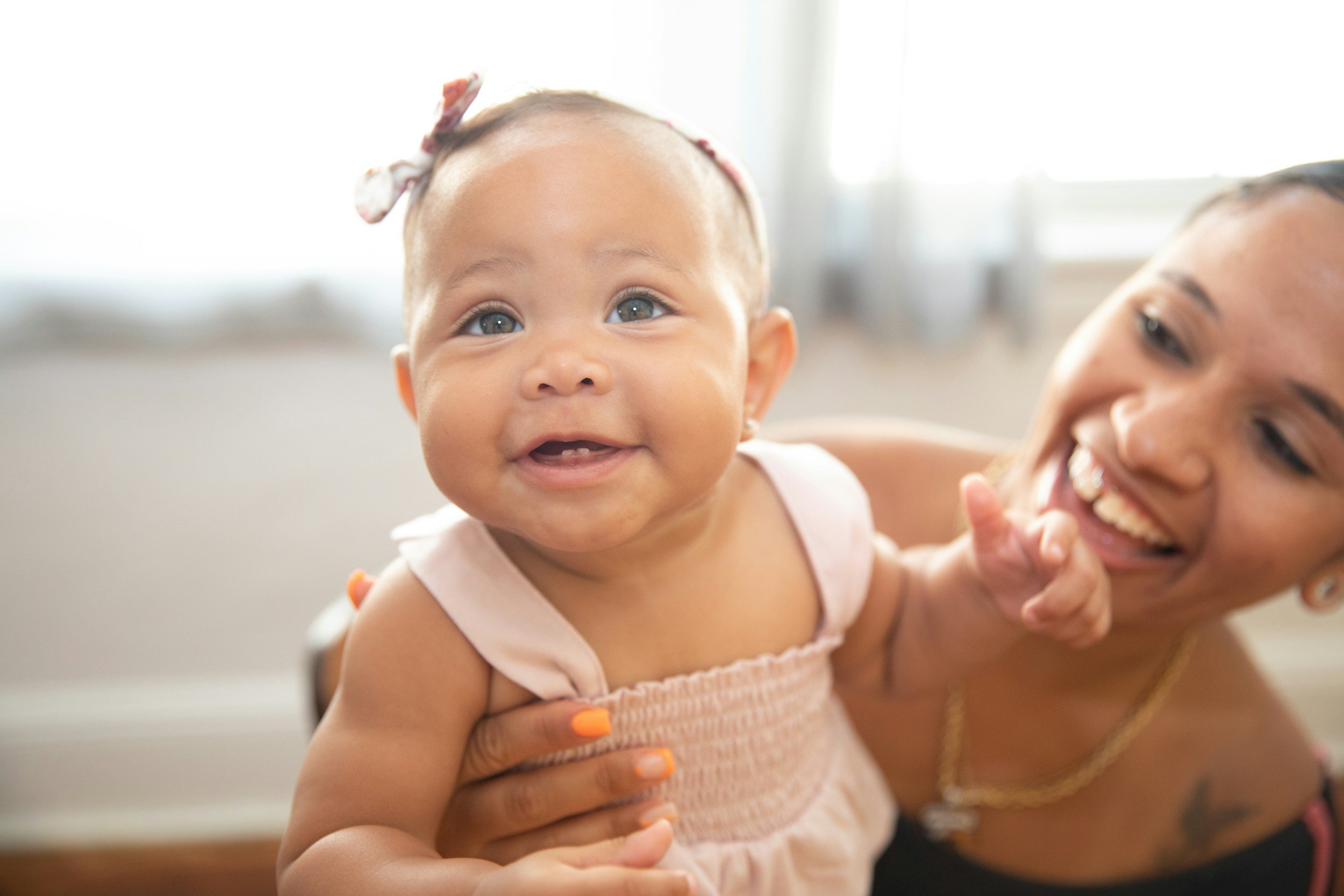 A smiling baby girl with blue eyes and a pink dress, holding hands with a woman with dark hair and a big smile, in a bright room.