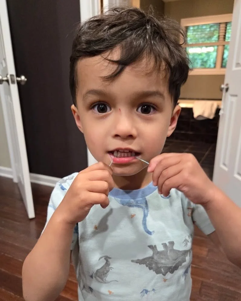 A young boy with dark hair and large eyes is holding a string in his mouth and looking directly at the camera. He is inside a house near an open door, with wooden flooring and a window in the background.