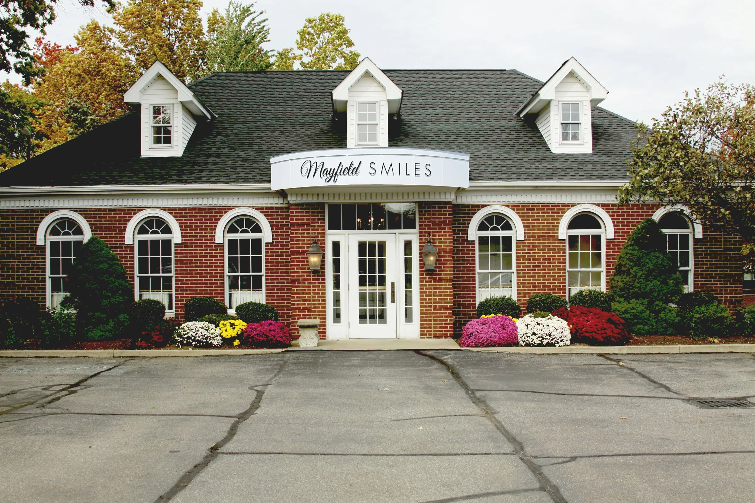 A brick building with a sign that reads "Mayfield Smiles" above the entrance, decorated with flower bushes and surrounded by a parking lot.