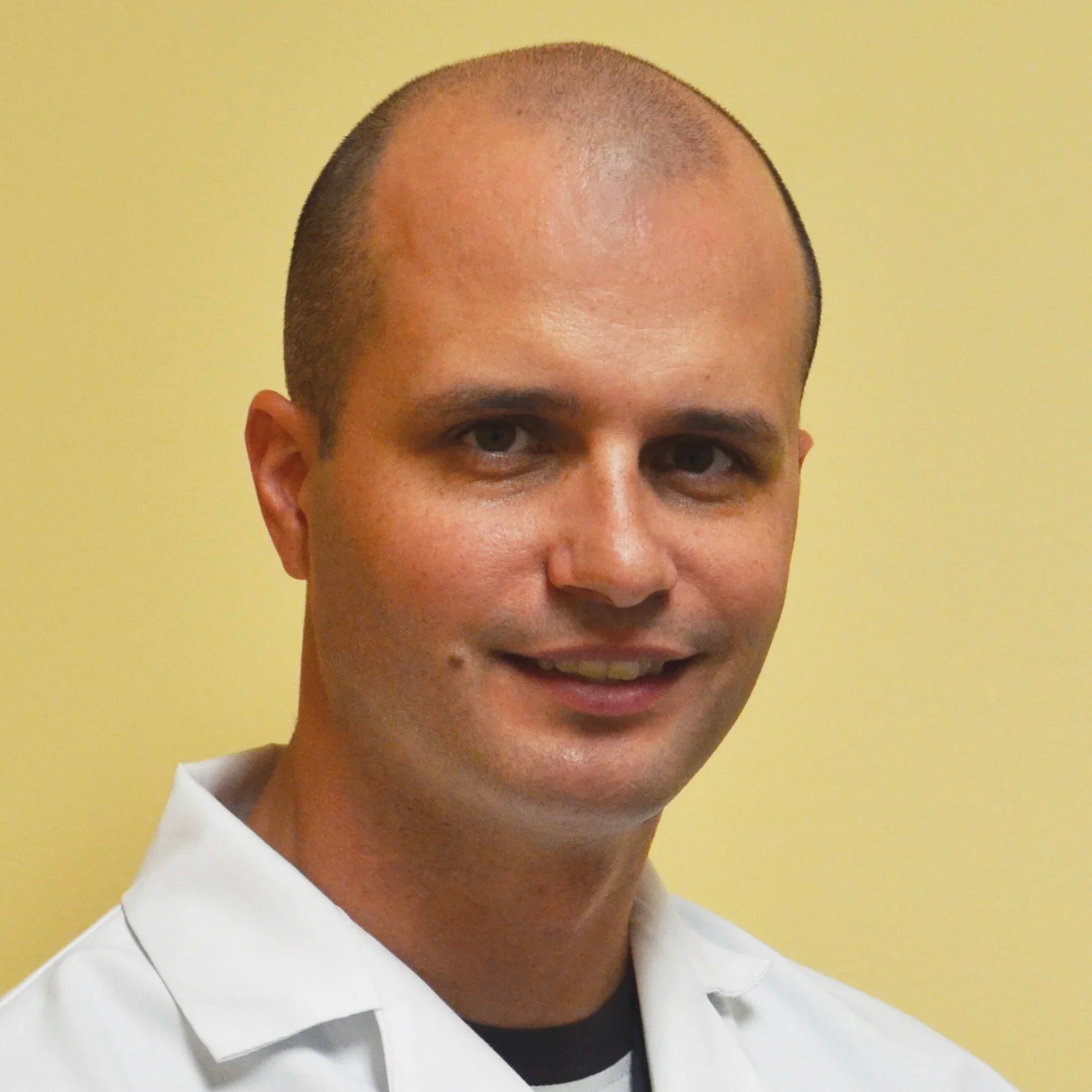 A portrait of a man with short, shaved hair, wearing a white collared shirt, standing against a yellow background.