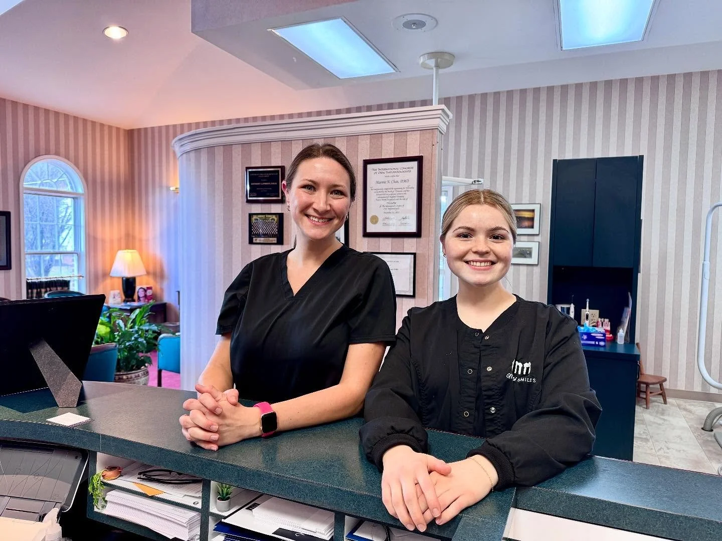 Two women smiling at a reception desk inside a dental office, with framed certificates on the wall behind them and dental equipment on a counter to the right.