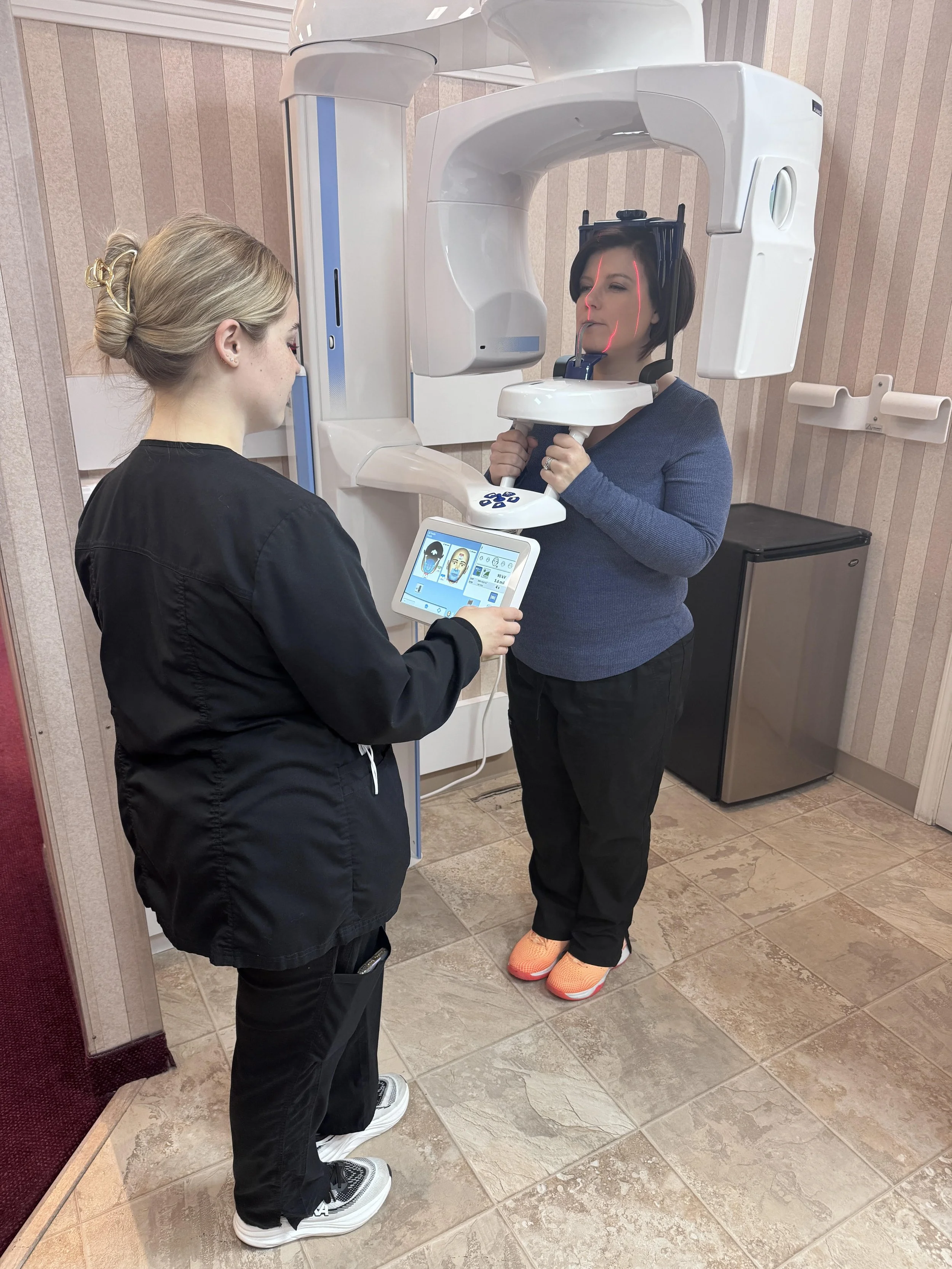 A woman undergoing a dental scan uses a panoramic X-ray machine while a technician monitors the process from a tablet.