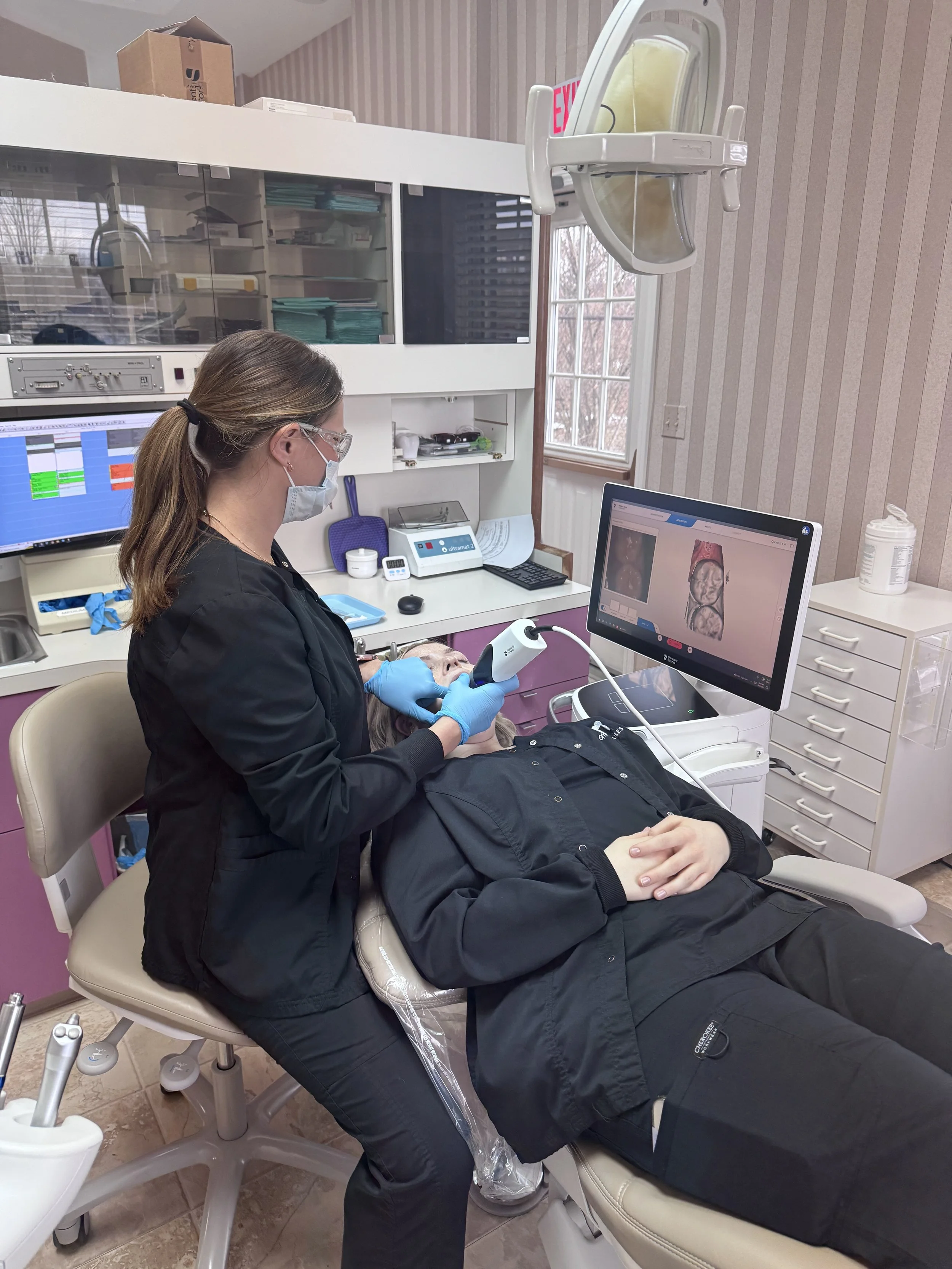 A medical professional performing a dental or oral health scan on a patient using a handheld device, with a monitor displaying a 3D scan of the patient's mouth or teeth in a dental clinic.