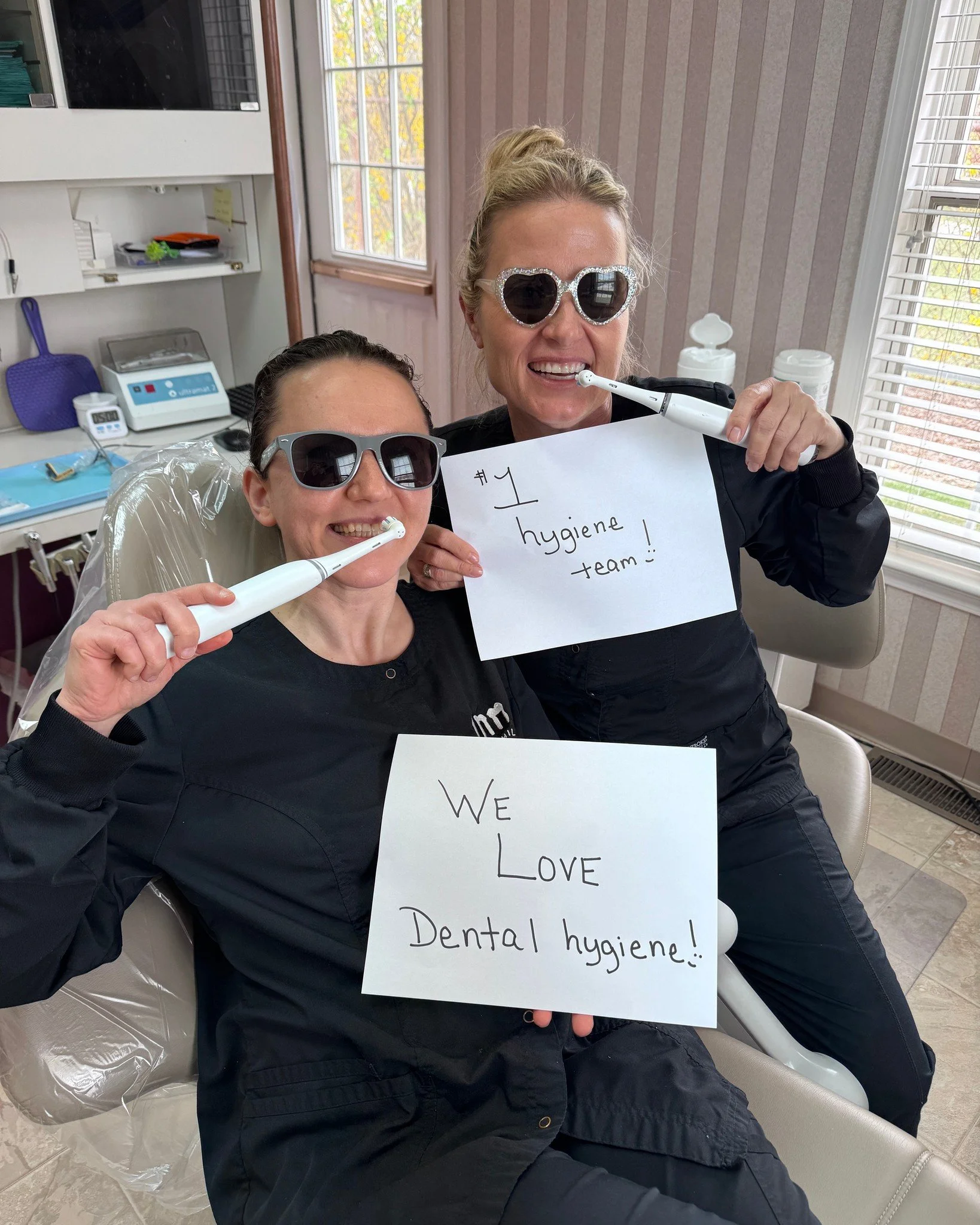 Two women wearing sunglasses holding dental tools and signs promoting dental hygiene in a dental office.