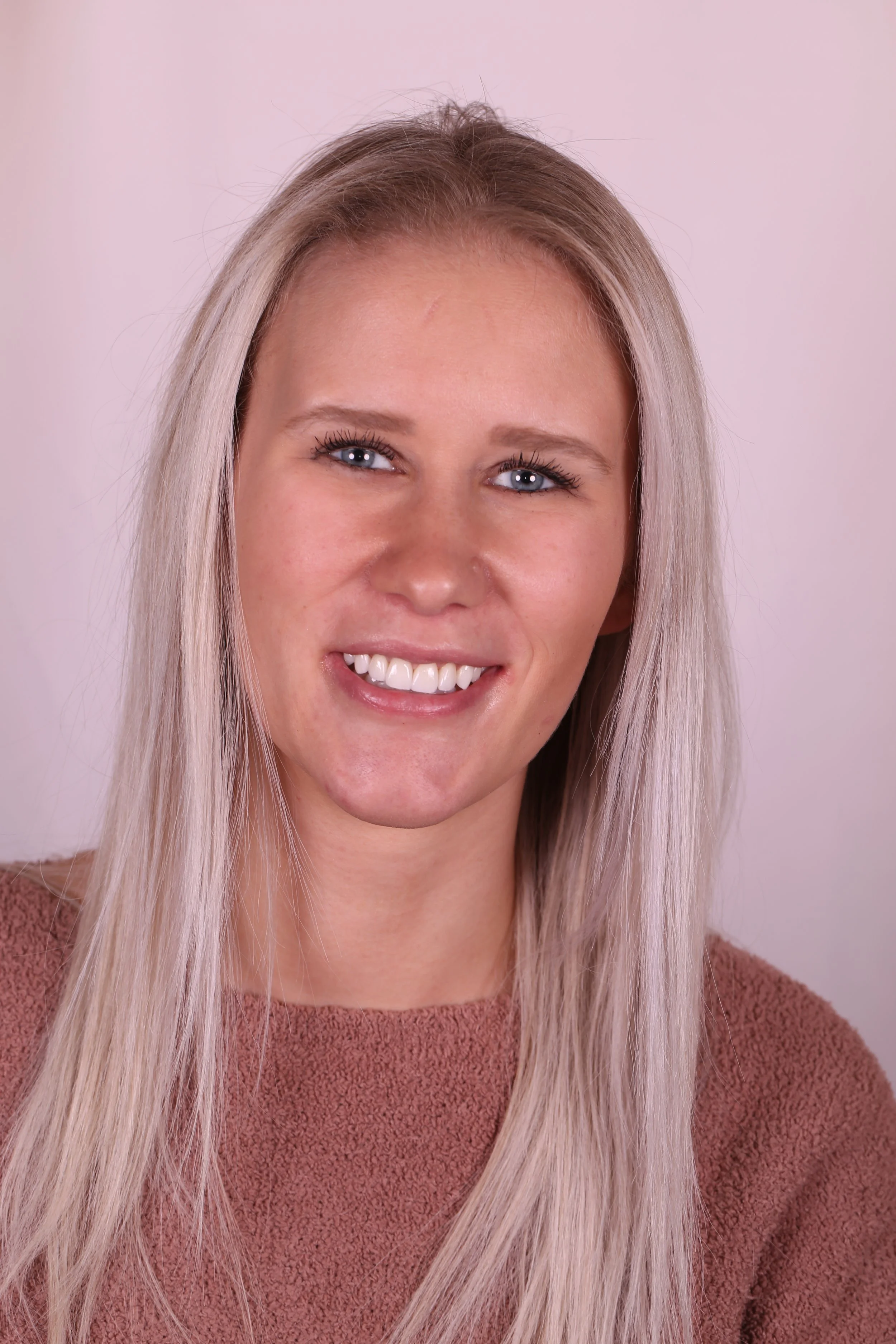 A smiling woman with long blonde hair and blue eyes, wearing a brown top, against a light pink background.