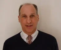 A man wearing a dark sweater over a collared shirt with a patterned tie, standing against a plain wall.