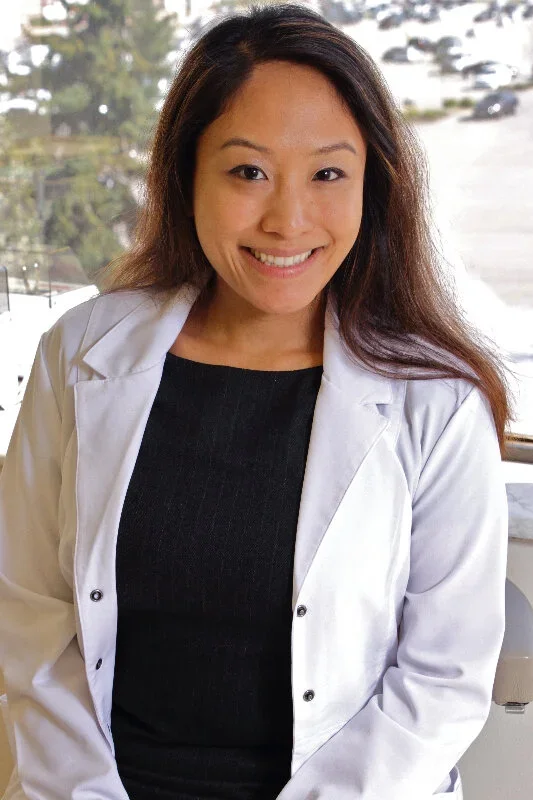 A woman with long dark hair wearing a black top and white coat, smiling, standing in front of a window with trees and cars outside.