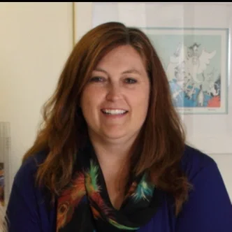 Smiling woman with shoulder-length brown hair, wearing a colorful scarf and blue top, standing in front of a wall with framed artwork.