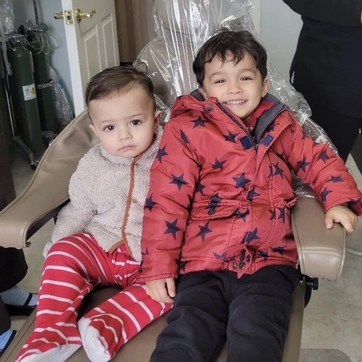 Two young boys sitting on a brown dental chair, one in a red star pattern jacket and black pants, smiling, and the other in a cream fuzzy jacket and red striped pants, looking serious. The background shows a door, some oxygen tanks, and a person standing nearby.