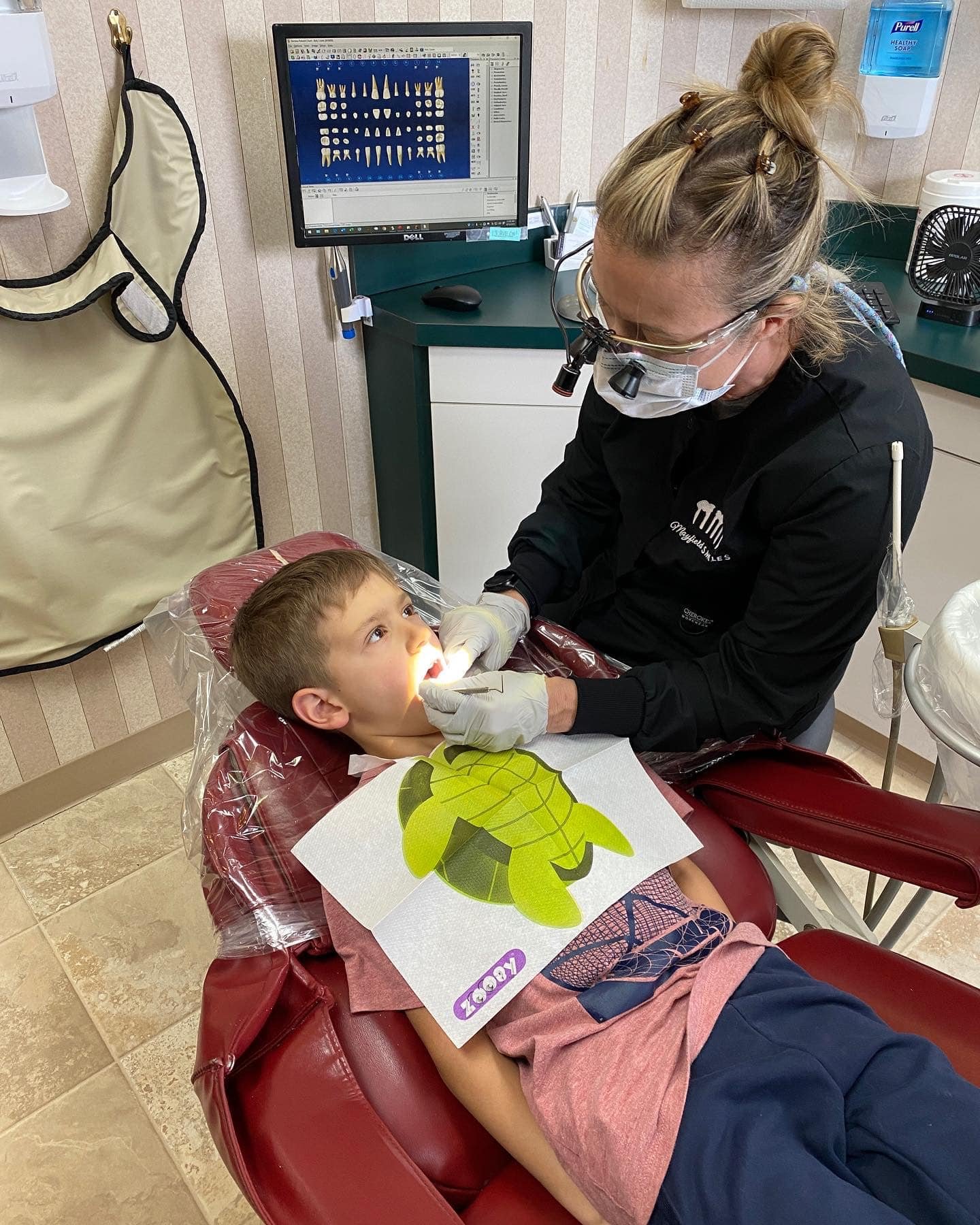 A young boy getting a dental checkup from a female dentist in a dental office. The boy is lying back in a dental chair with a protective bib, and the dentist is examining his mouth with tools. A dental printout of leaves is on the boy's chest. In the background, there is a computer monitor displaying a diagram of teeth, a sanitizer dispenser, and other dental equipment.