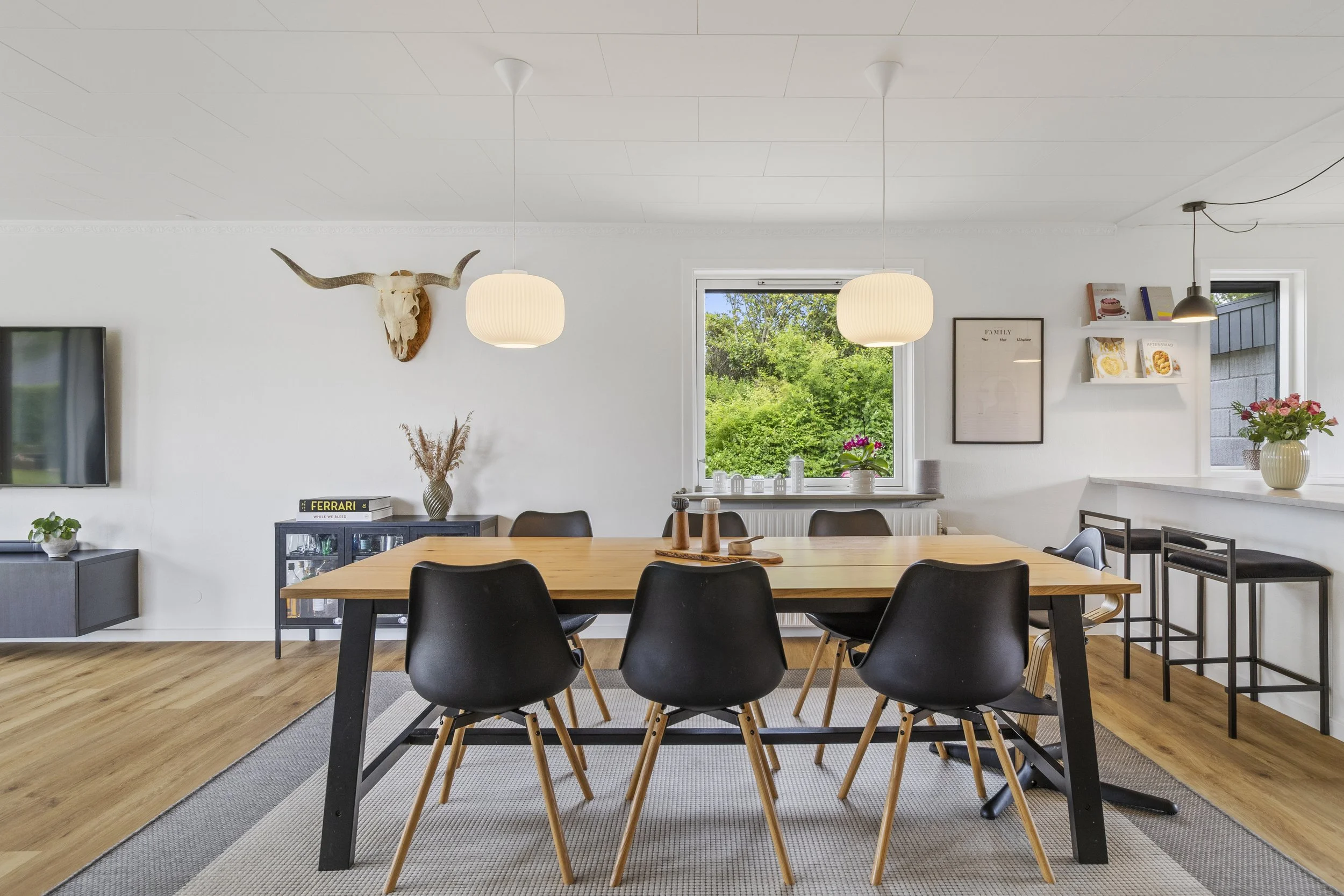 Moderne spisestue med træbord, seks sorte stole, to hængelamper, vindue mod grønt udeområde samt dekorative genstande og opsat dyrekranium.