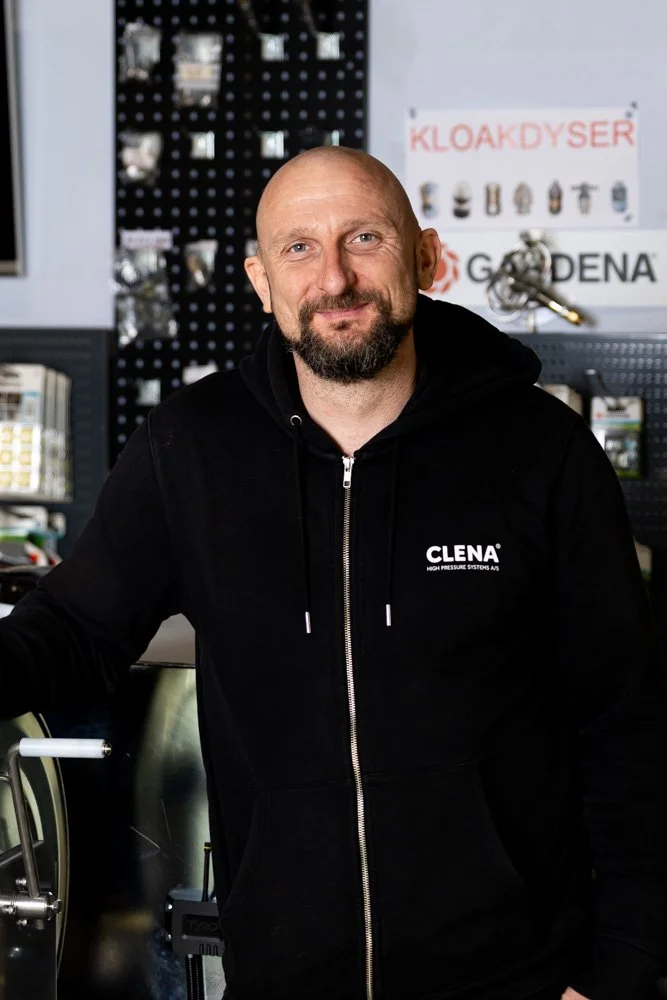 Skaldet mand med skæg i sort hoodie med ’CLENA’-logo, stående i butik med værktøj og maskiner i baggrunden.