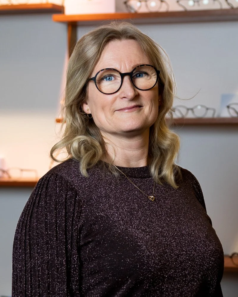 Kvinde med blond hår og briller i sort glitrende top, indendørs foran hylder med briller.