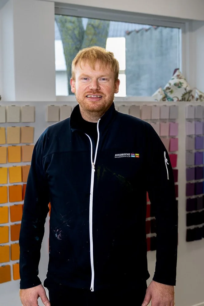 Mand med rødt hår og skæg, smilende, iført sort jakke med farvet logo foran væg med malingsprøver.