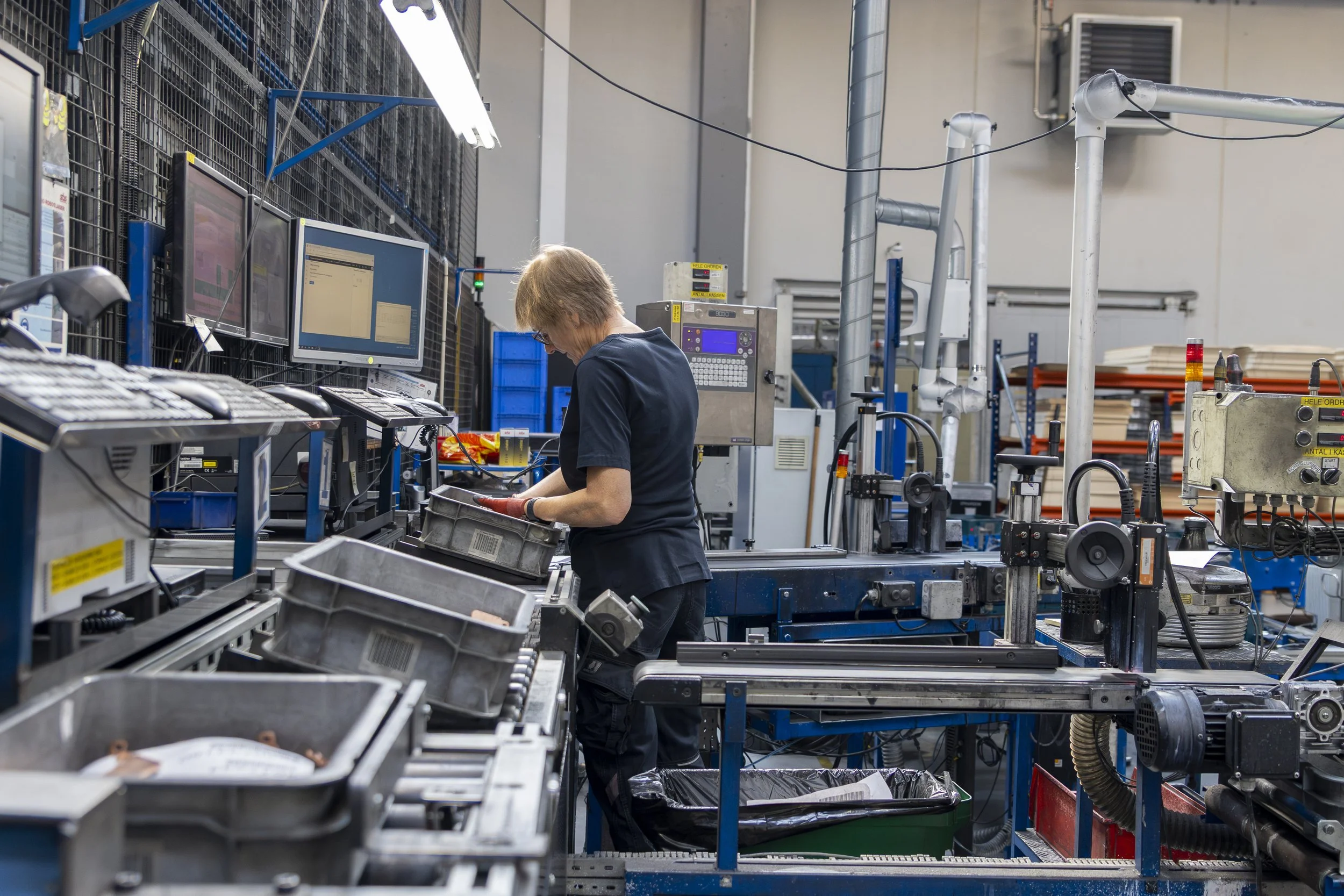 Arbejder i industri- eller produktionsmiljø ved arbejdsbord, sorterer eller inspicerer dele med maskiner og skærme i baggrunden.