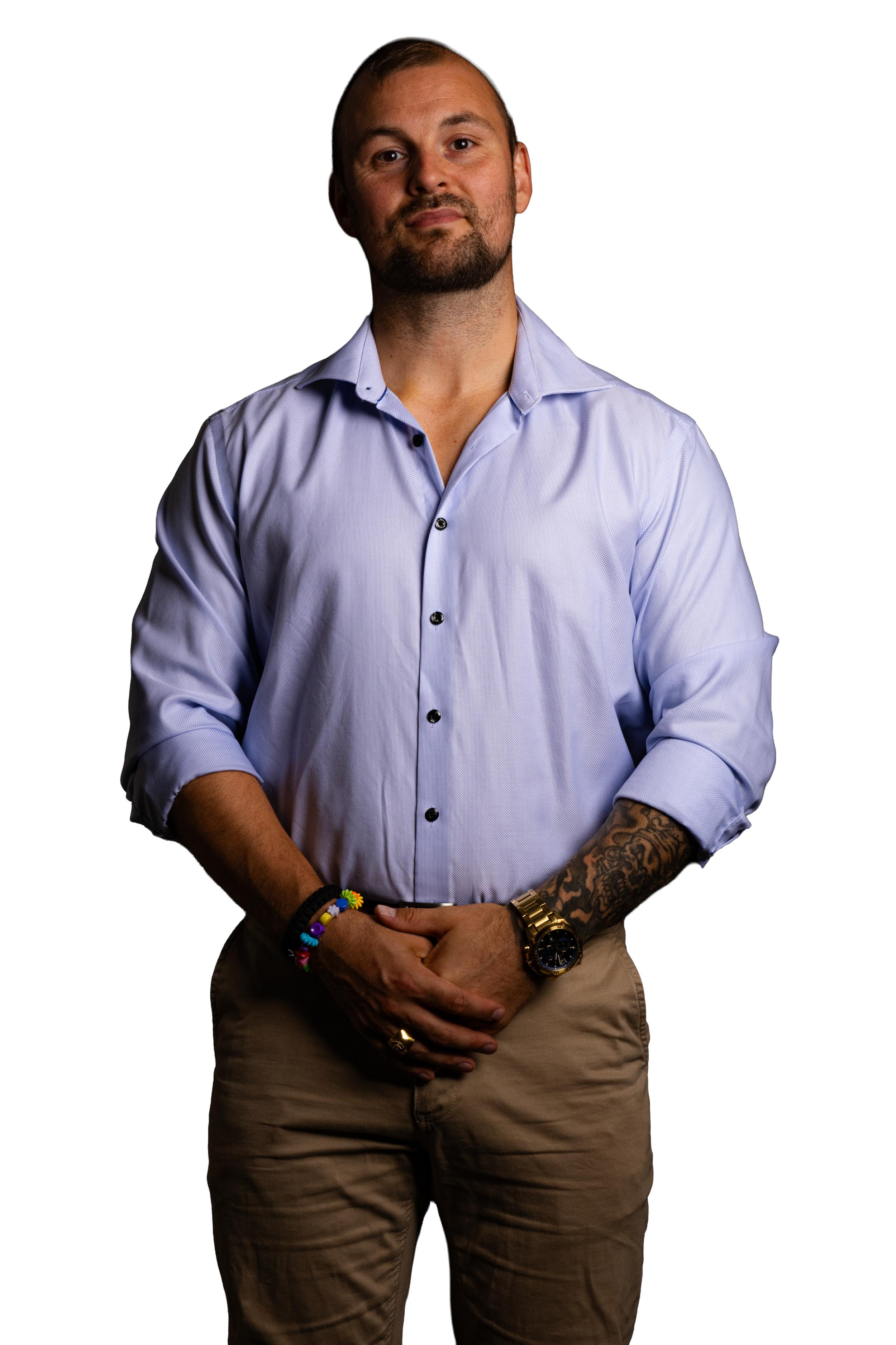 Mand med skæg i lyseblå skjorte med oprullede ærmer, khakibukser, guldur og farverige armbånd, stående mod sort baggrund.