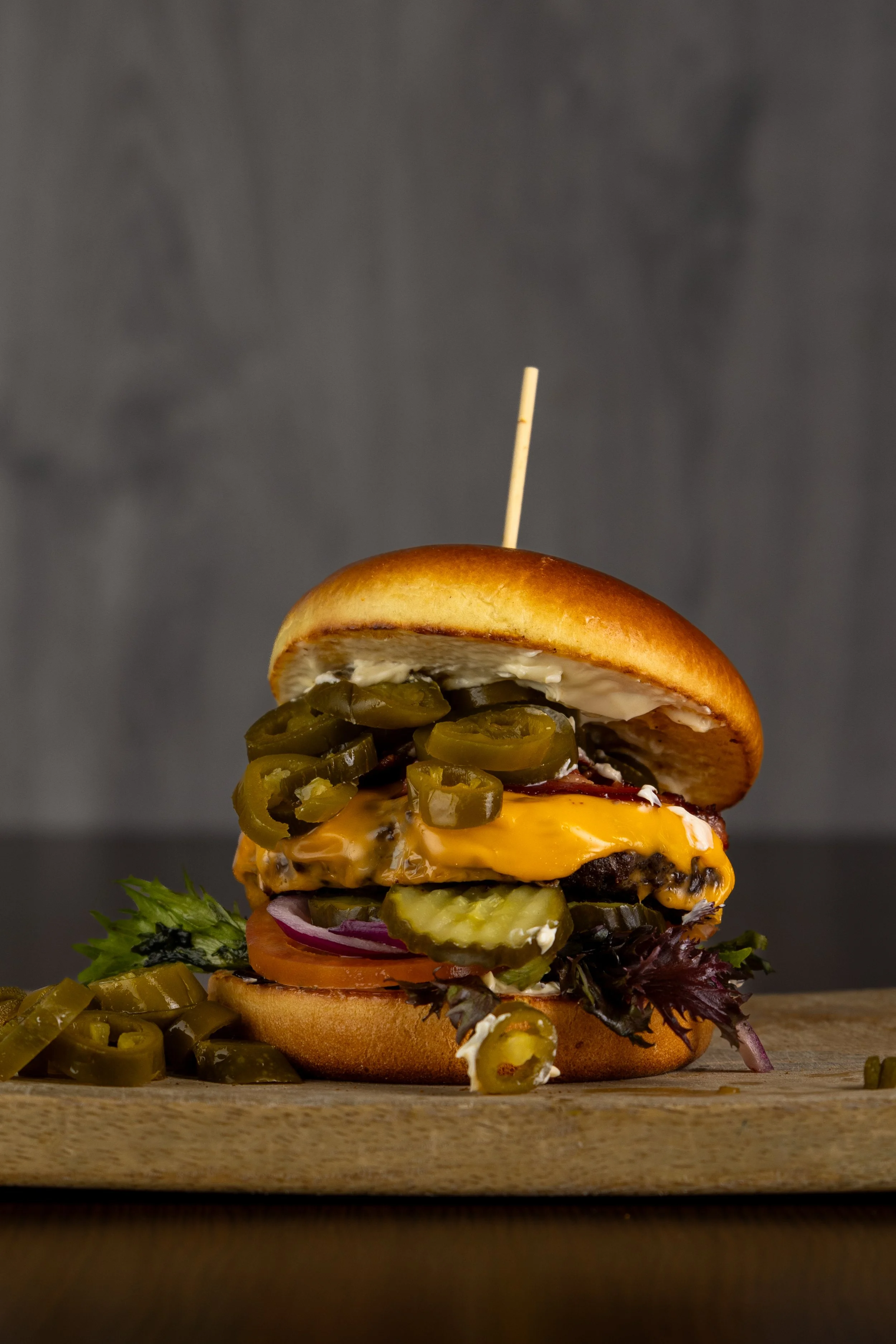 Close-up af cheeseburger med pickles, agurk, tomat, rødløg, salat, smeltet ost og bøf i bolle, med tandstik i toppen på træbord.