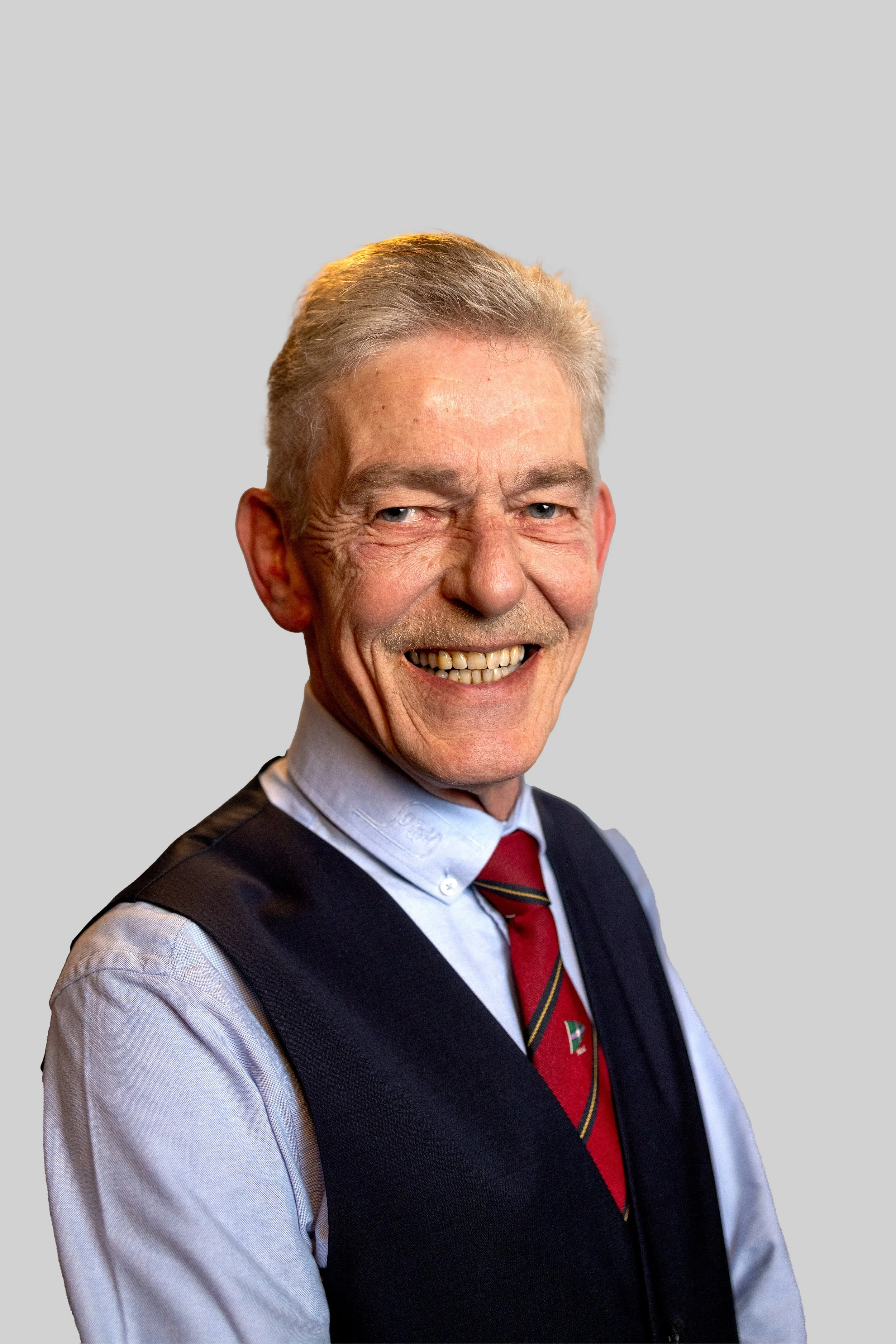 Ældre smilende mand med gråt hår, lyseblå skjorte, rød mønstret slips og mørk vest mod neutral baggrund.