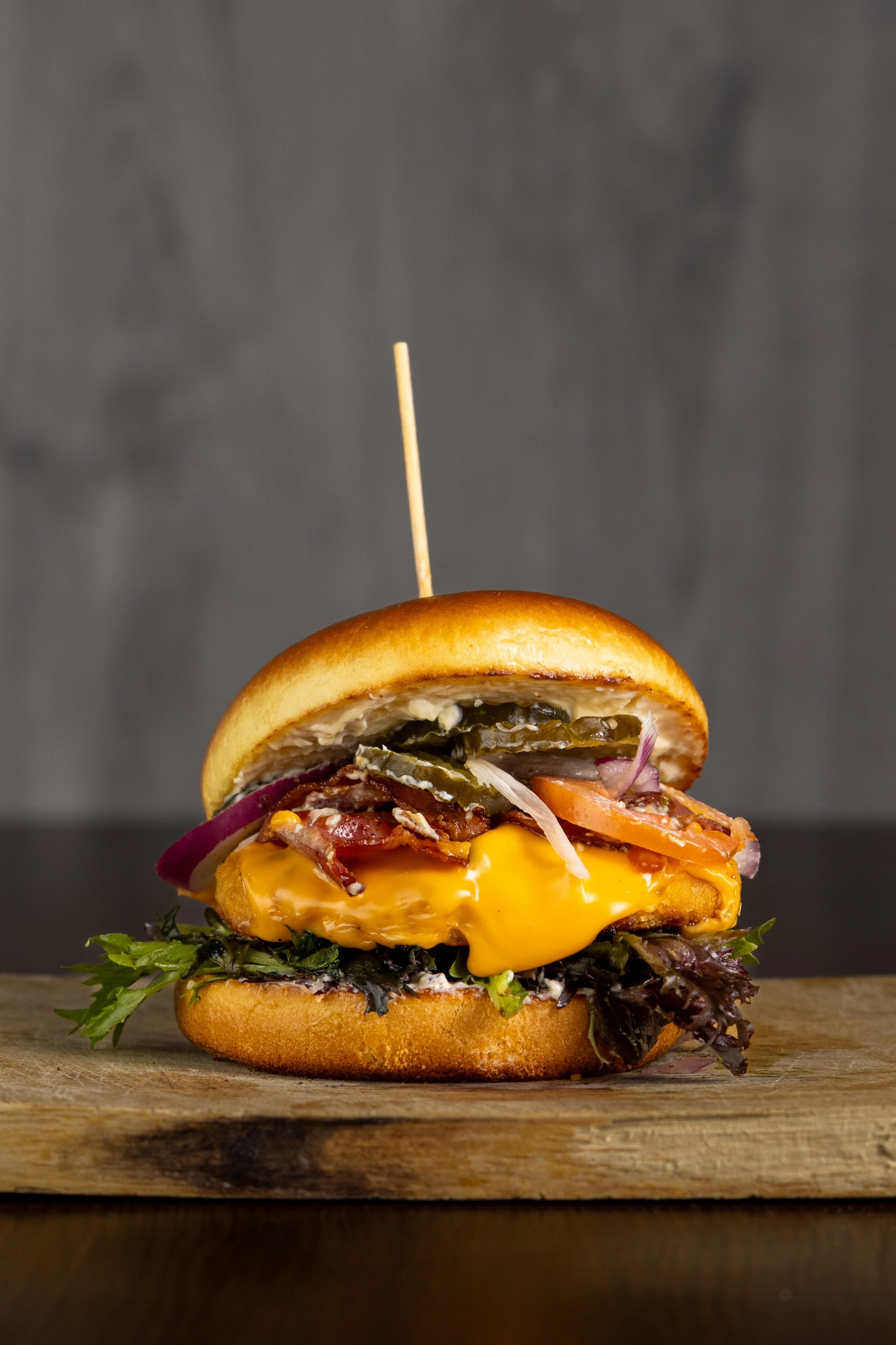 Close-up af cheeseburger med pickles, bacon, løg og salat på træoverflade.