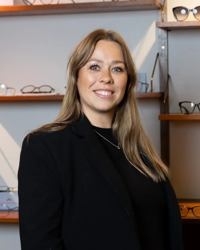 Kvinde med langt blond hår i sort blazer og halskæde, stående foran hylder med solbriller og briller.