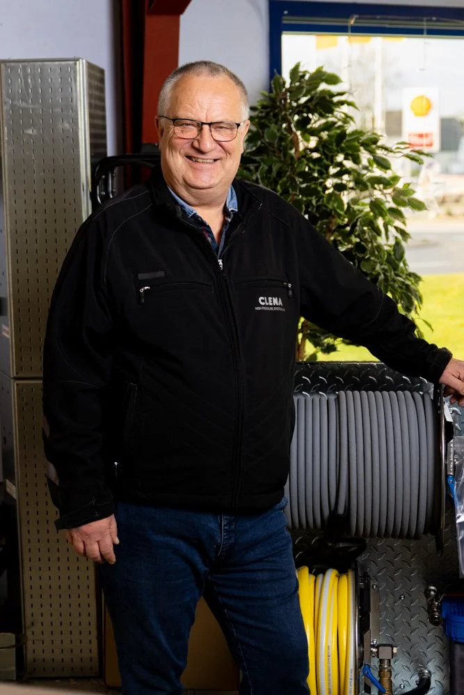 Smilende mand med briller og sort jakke med ’Clena’-logo, indendørs i butik med stort grønt plante og slanger/udstyr bagved.