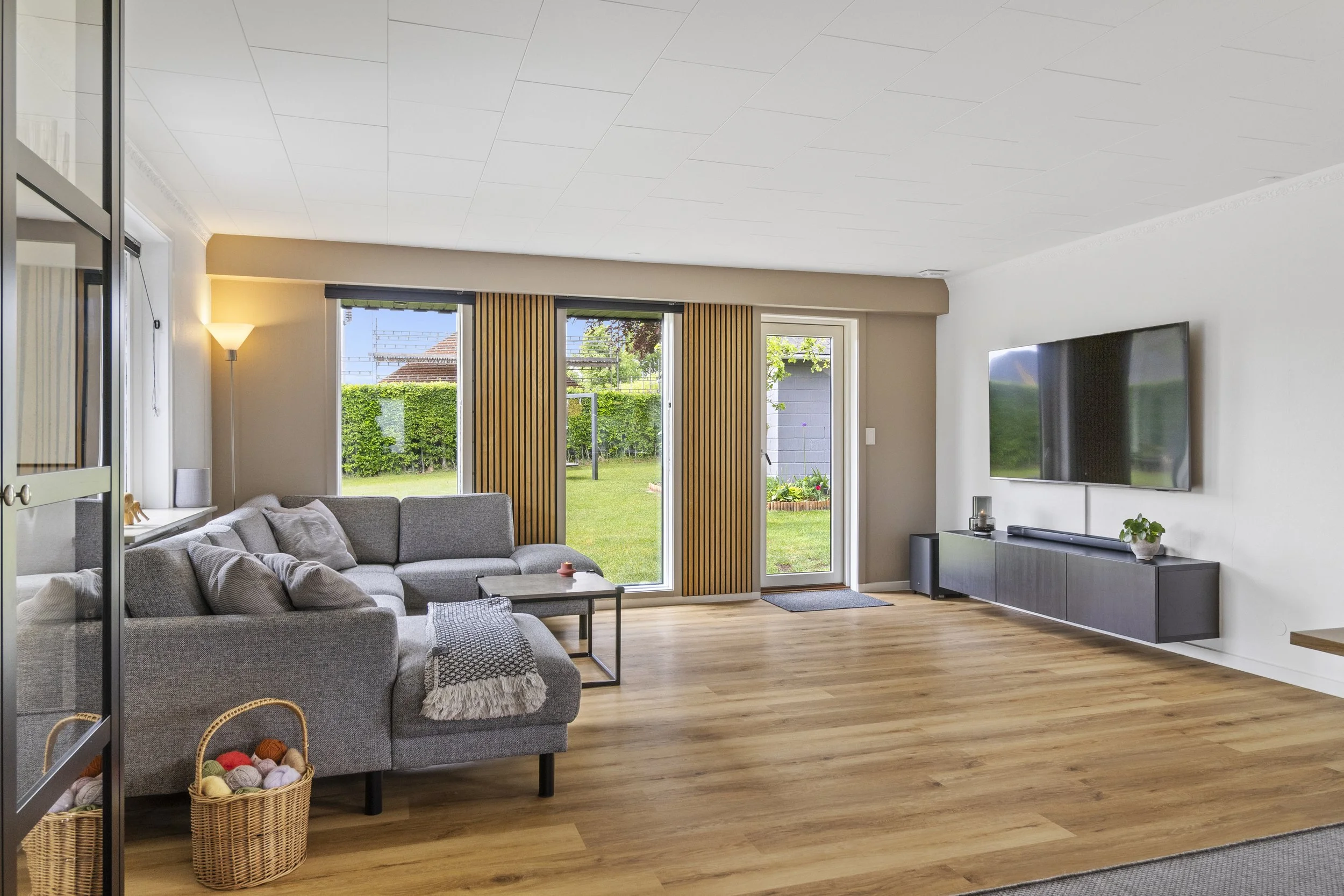 Stue med grå hjørnesofa, kurv med garn, træ-sofabord og skydedøre til grøn have.