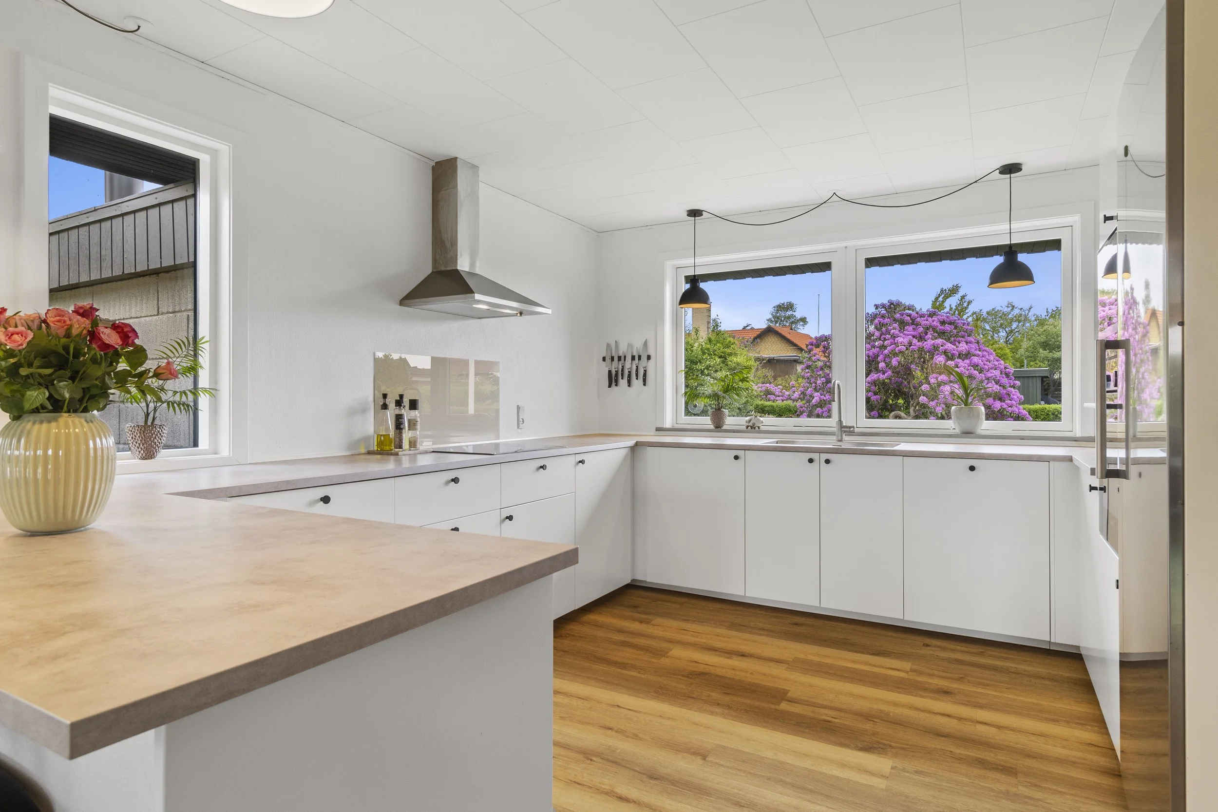 Lys køkken med hvide skabe, træbordplade, store vinduer mod have med lilla blomster og sorte pendellamper, vase med lyserøde blomster på bordet.