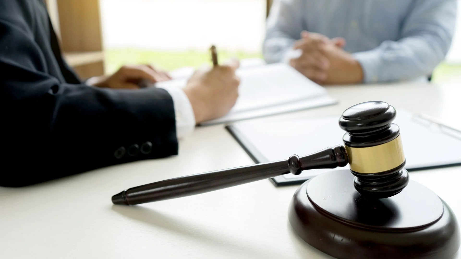 A judge's gavel rests on a white table in a courtroom, with two people sitting across from each other in the background, one taking notes.