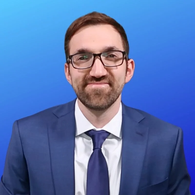A man with glasses, groomed beard, wearing a blue suit, white shirt, and blue tie, smiling in front of a blue gradient background.