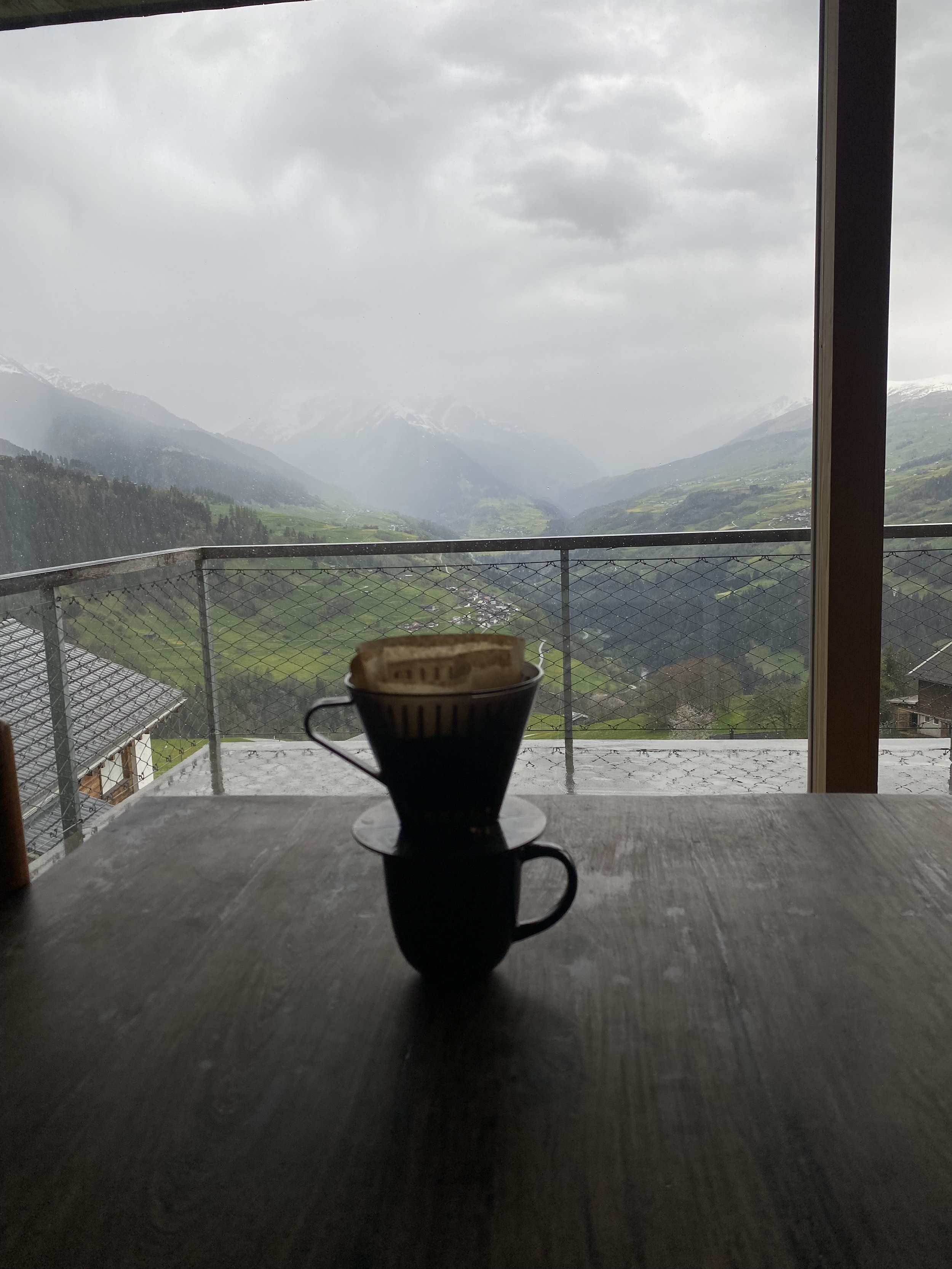 A cup of coffee with a slice of bread on top, placed on a dark wooden table, overlooking a misty mountain landscape with green valleys and snow-capped peaks.
