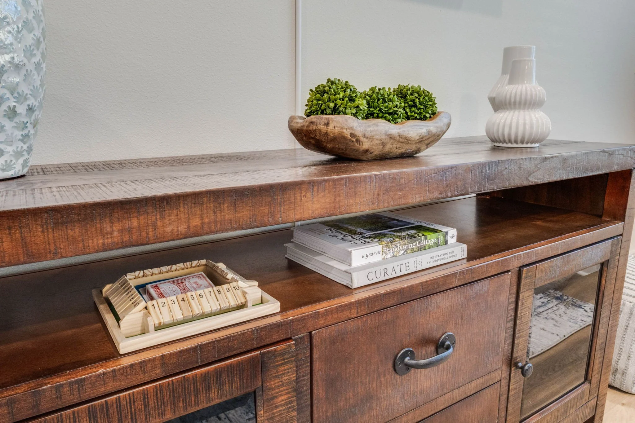 Wooden sideboard with decorative items including a small potted plant, a white ceramic vase, and a tray of small wooden dominoes, with a stack of books underneath.