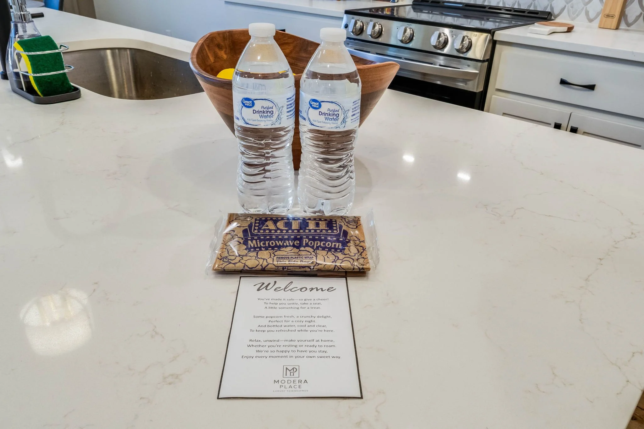 Two bottles of bottled drinking water, a packet of microwave popcorn, and a Welcome note on a white kitchen countertop.