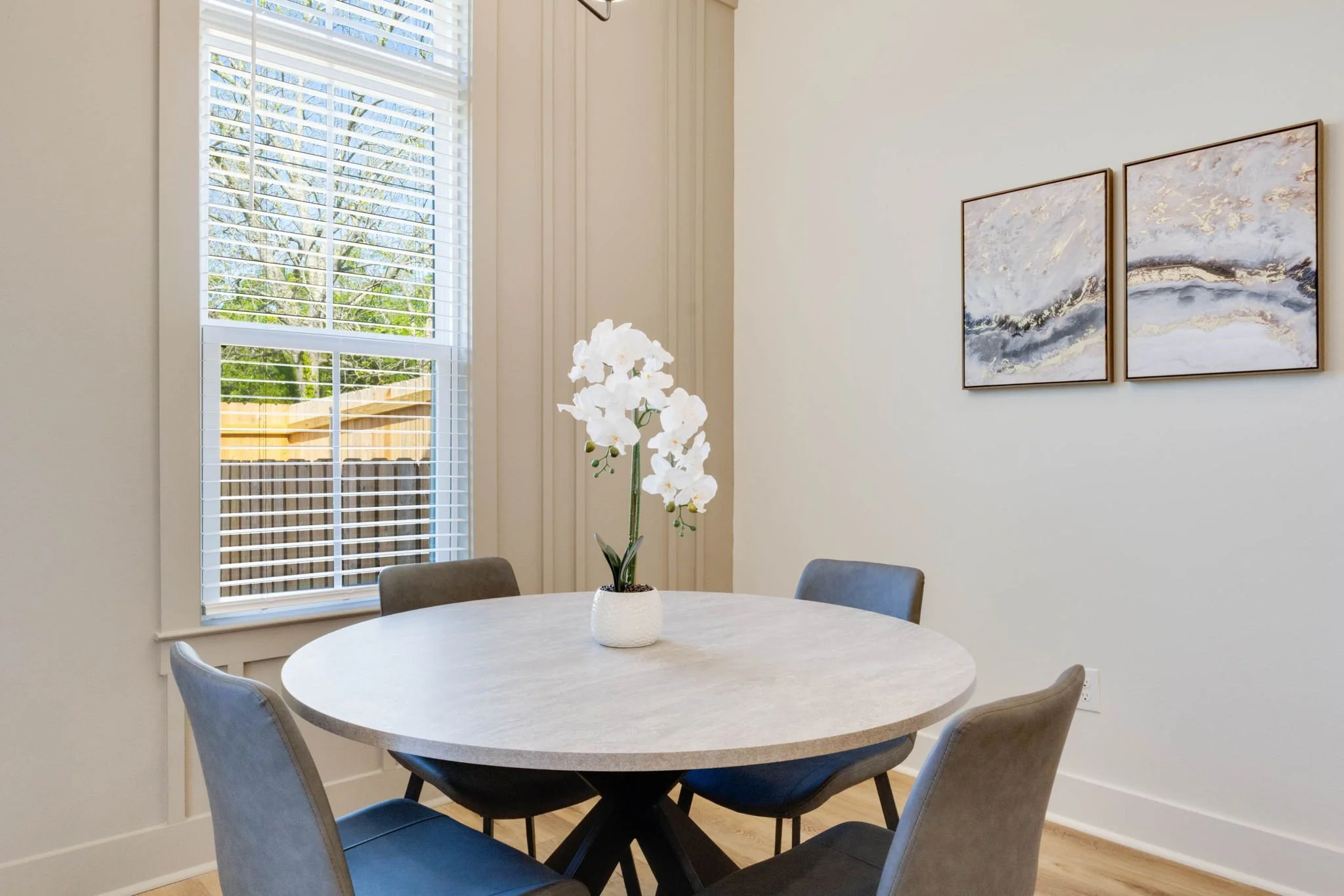 A dining room with a round table, four chairs, a white orchid in a white vase, a window with white blinds, and two abstract paintings on the wall.