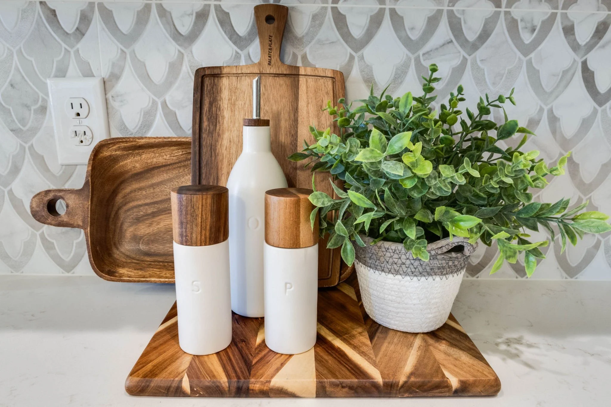 Decorative kitchen countertop with wooden trays, salt and pepper shakers, a white condiment bottle, a potted green plant, and a tiled wall background.