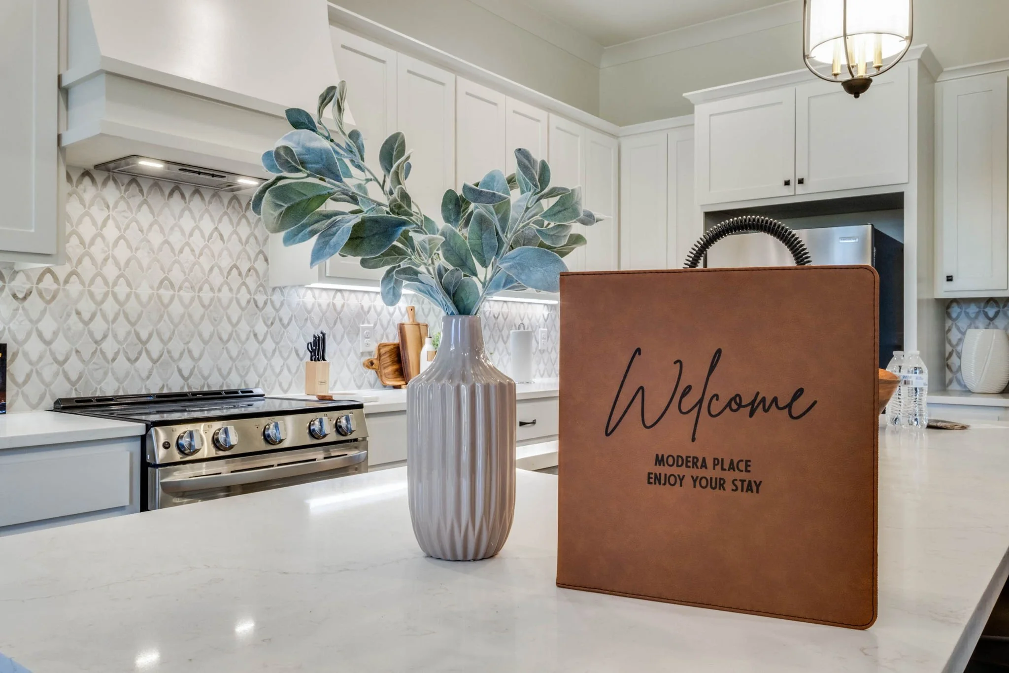 A modern kitchen with white cabinets, a stainless steel stove, and a white countertop. A white vase with green foliage and a welcome sign reading 'Welcome Modera Place Enjoy Your Stay' are on the counter.