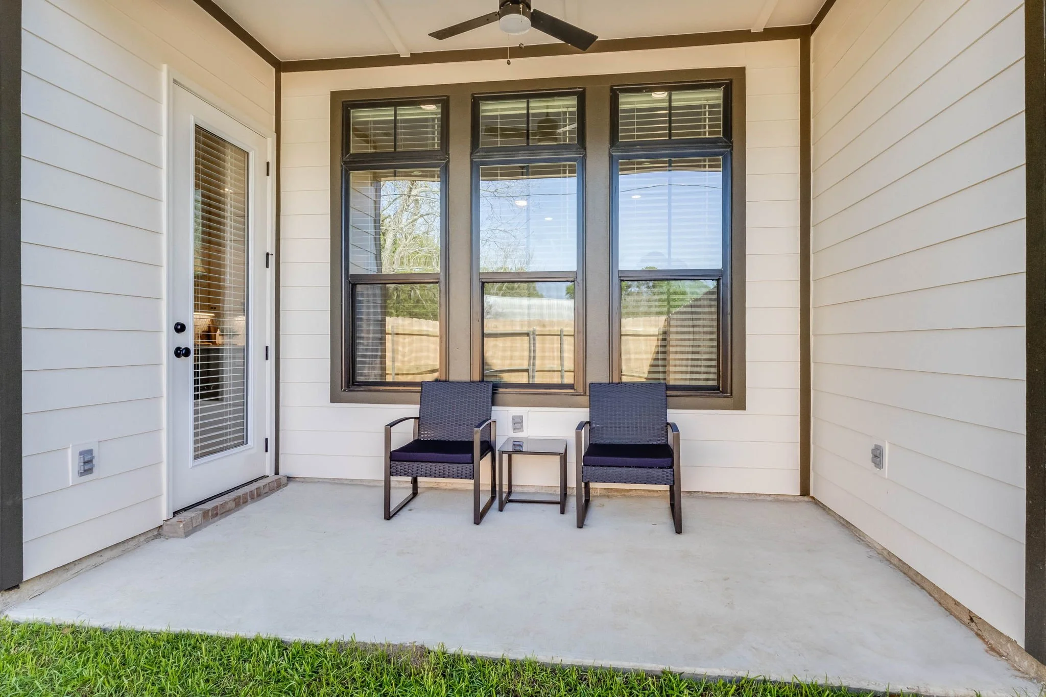 Back porch with two dark chairs and small table, large windows, white siding, ceiling fan, door with glass panel and blinds, green grass