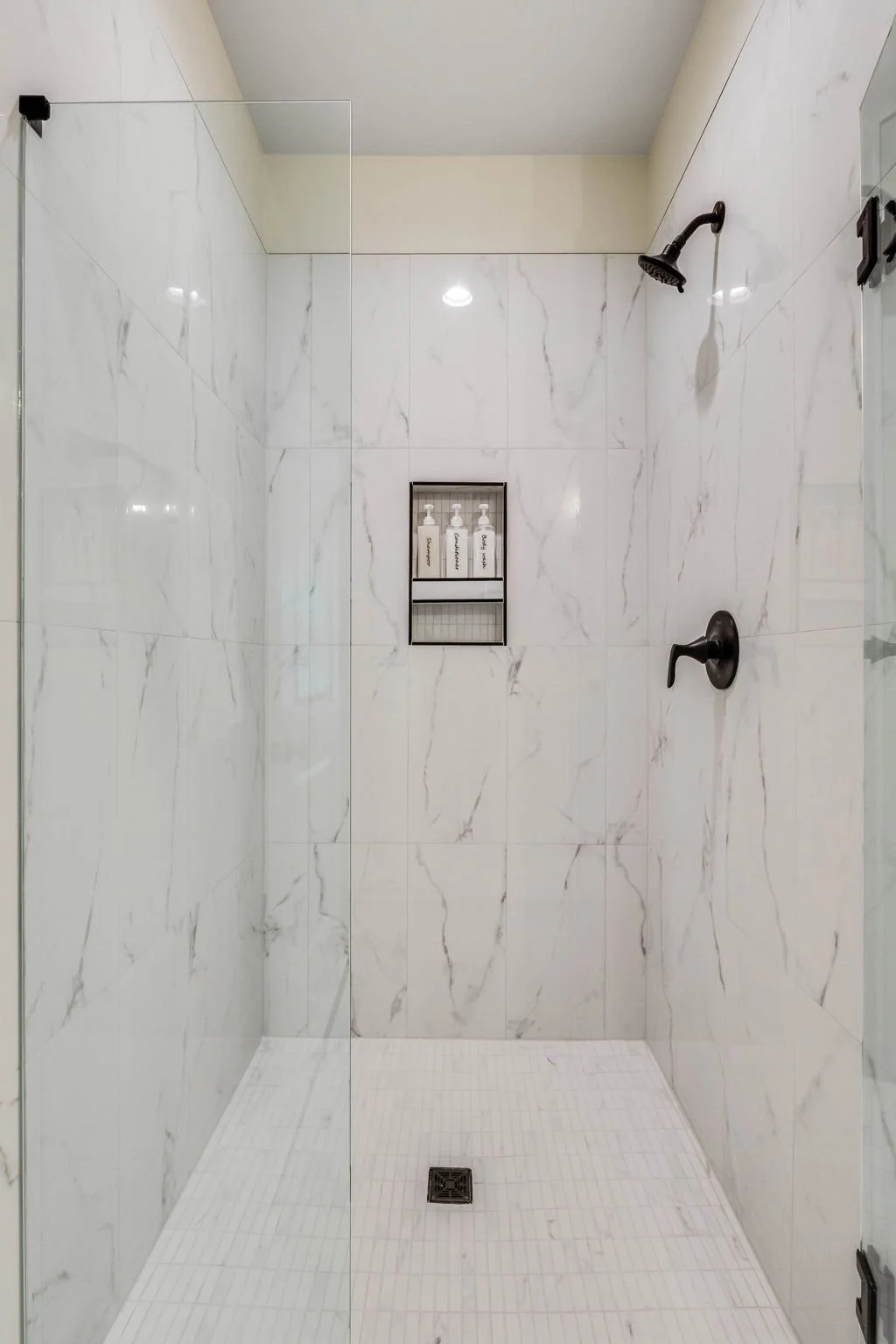 Empty walk-in shower with white marble tiles, a black showerhead and faucet, a built-in shelf with shower products, and a glass door.