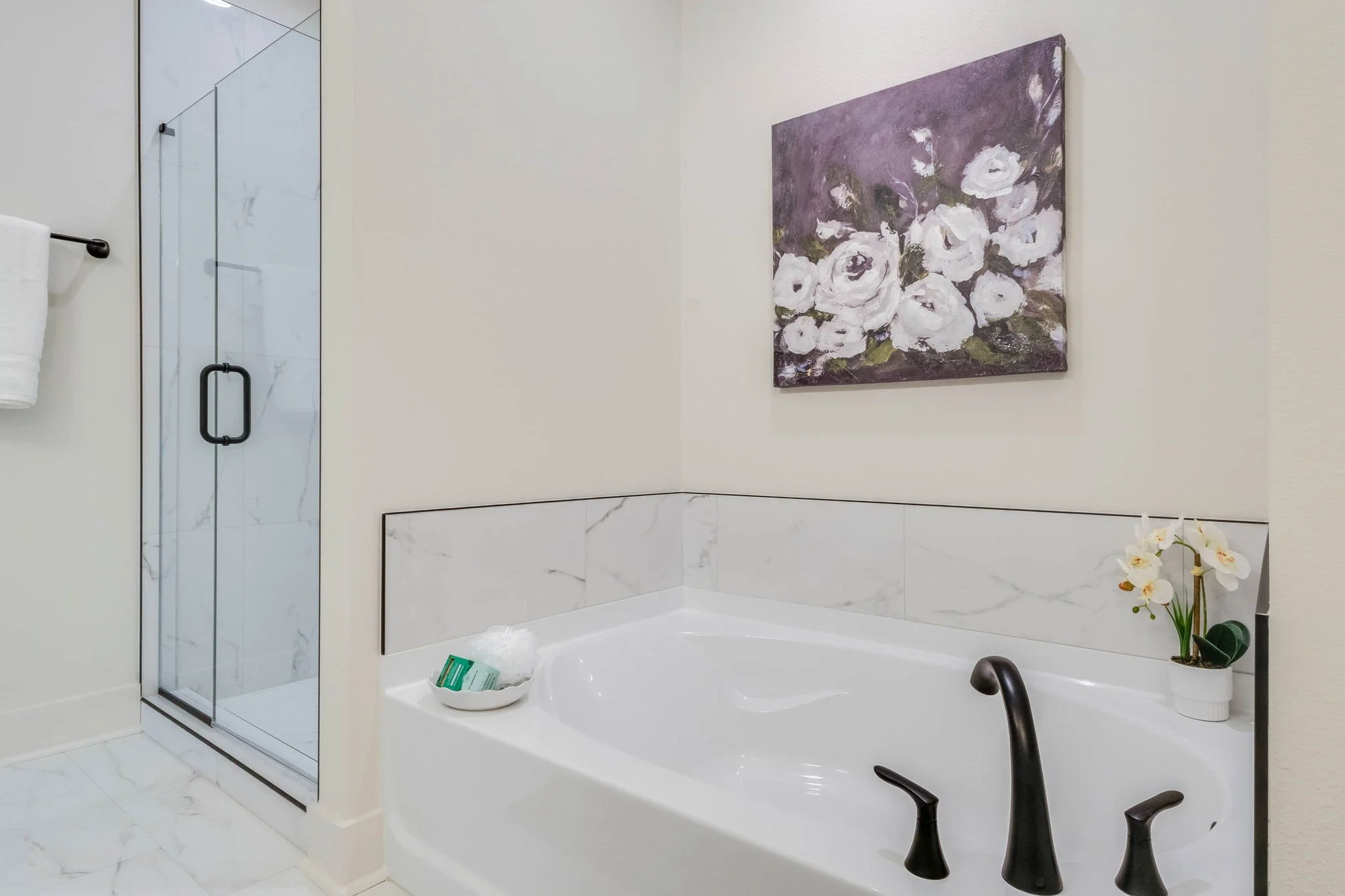 A bathroom corner with a white bathtub, a glass shower enclosure, a white orchid plant, a painting of white flowers on a purple background, and a rolled towel on the bathtub edge.