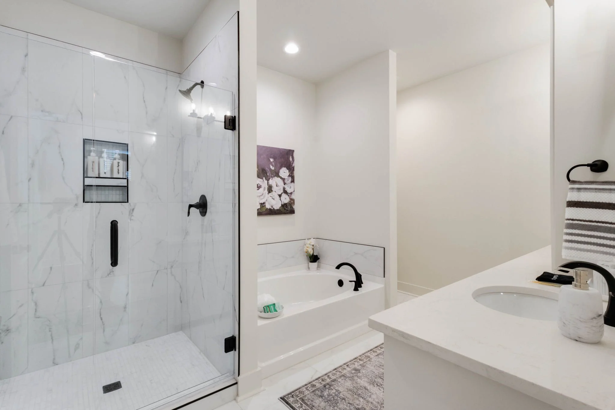 Modern bathroom featuring a glass shower with marble tiles, a bathtub with black fixtures, a white countertop with a round sink and soap dispenser, and a wall-mounted towel holder with a striped towel.