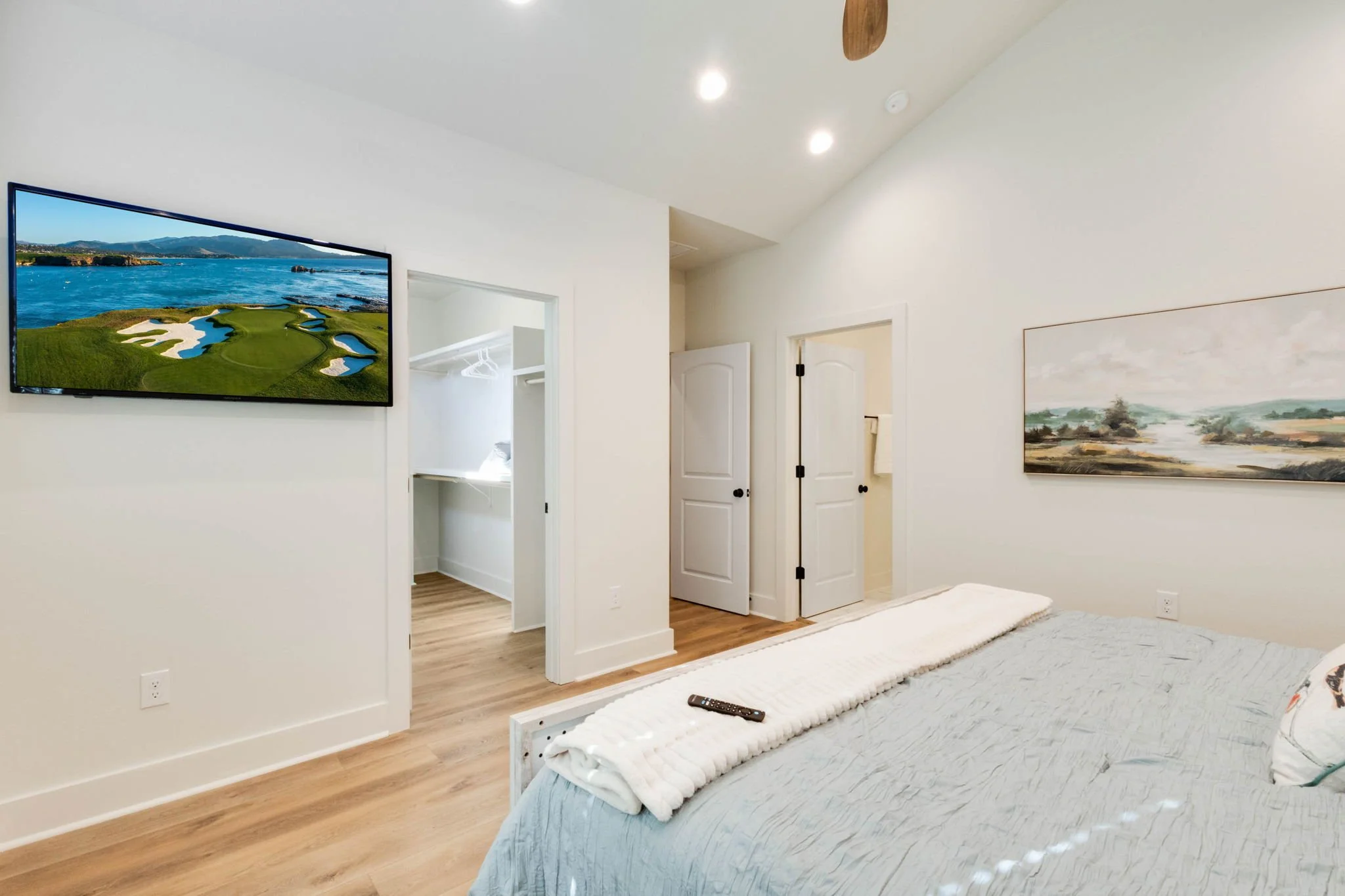 Modern bedroom with a TV showing a golf course, a bed with a gray bedspread and a white blanket, and a landscape painting on the wall.