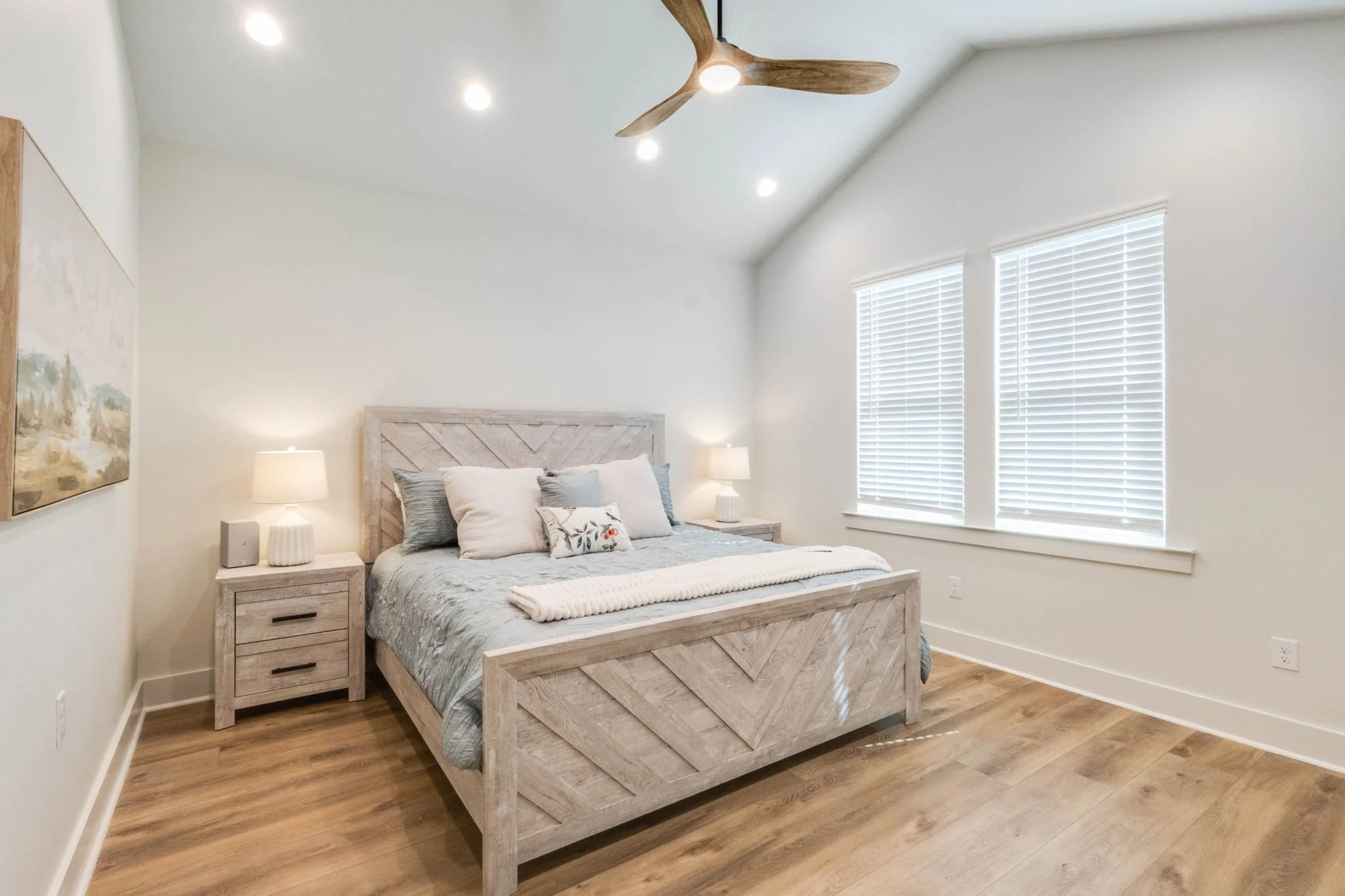 A bedroom with a wooden bed frame, matching bedside tables with lamps, a bed with a gray quilt and pillows, two windows with blinds, and a ceiling fan with wooden blades.
