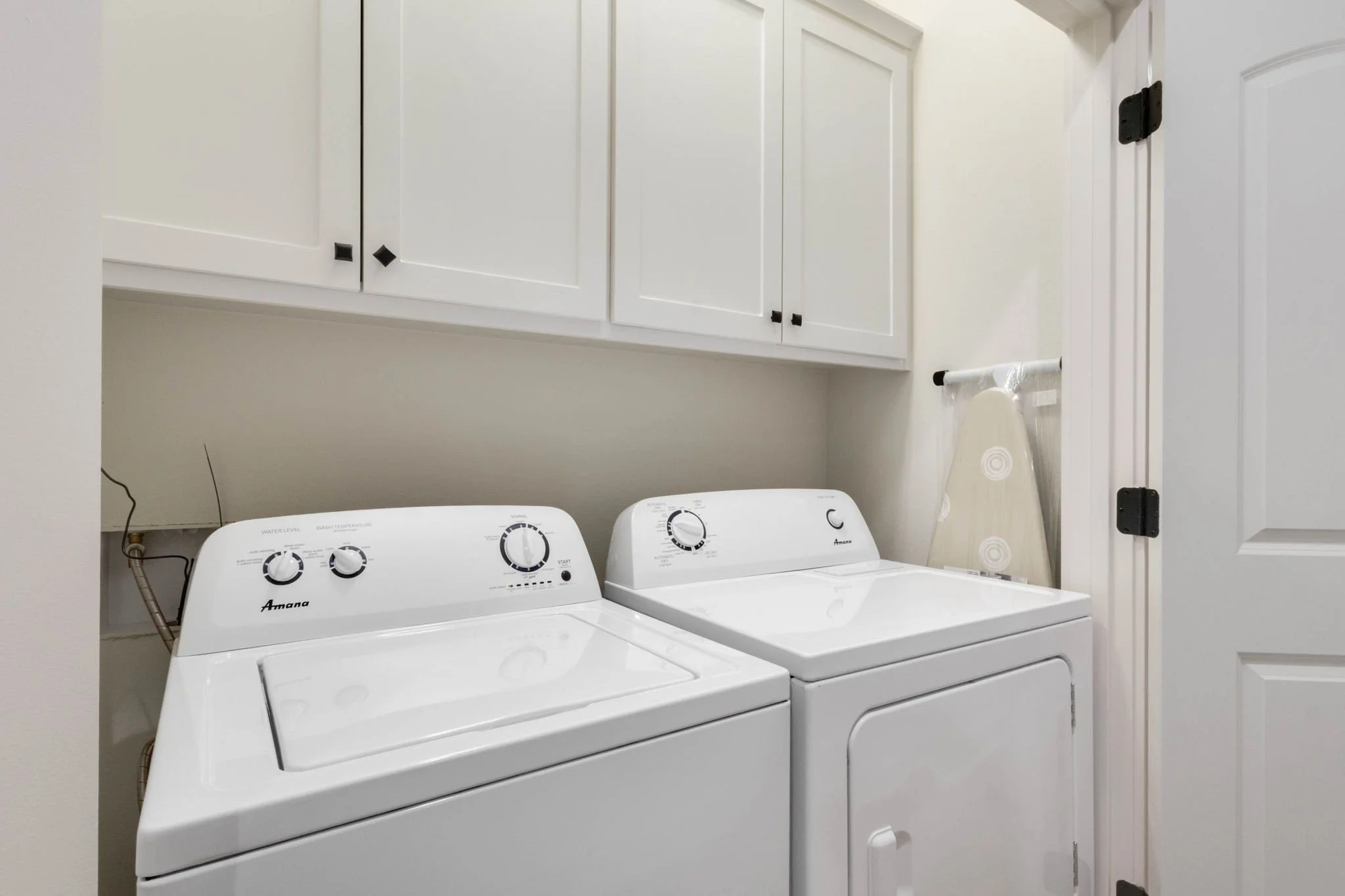 A laundry room with a white washer and dryer side by side, above which are white cabinets and an ironing board leaning against the wall.