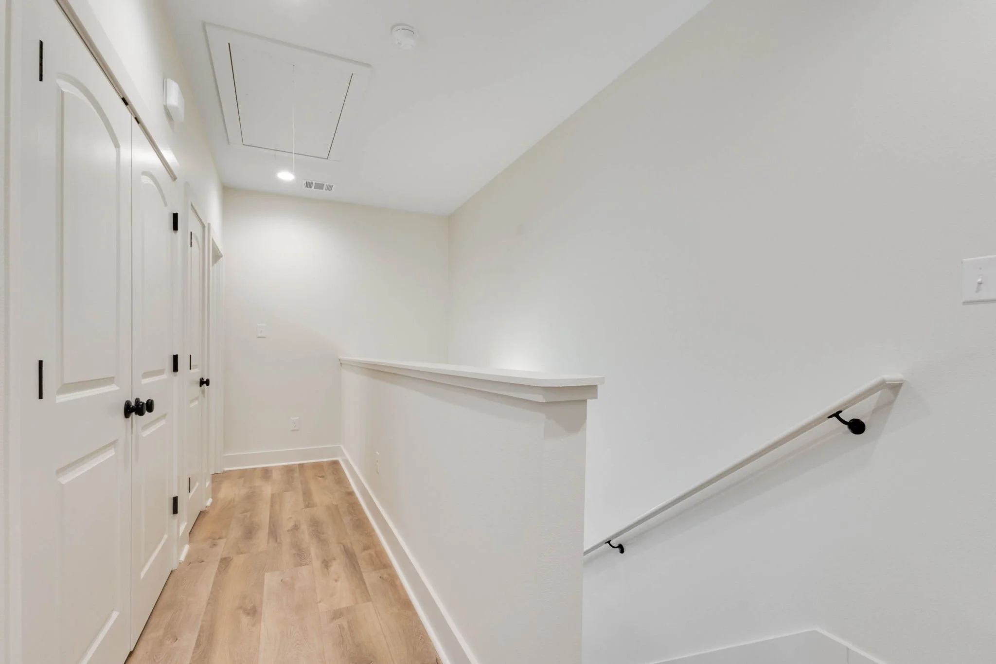 Interior view of a staircase landing with white walls, wooden flooring, closed white closet doors, a white handrail on the right, and ceiling lights.