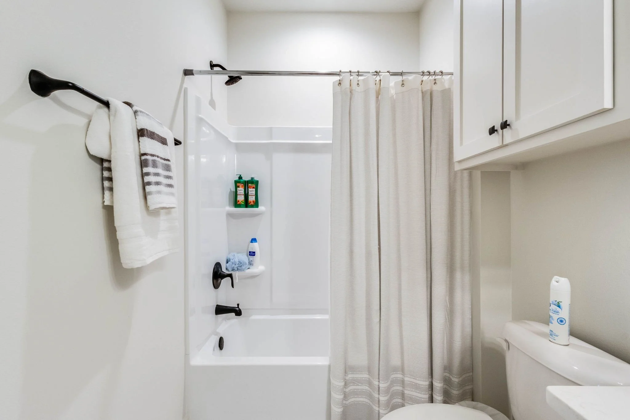 A clean, white bathroom with a bathtub and shower, beige curtains, wall-mounted cabinet, and towels hanging on a black hook.