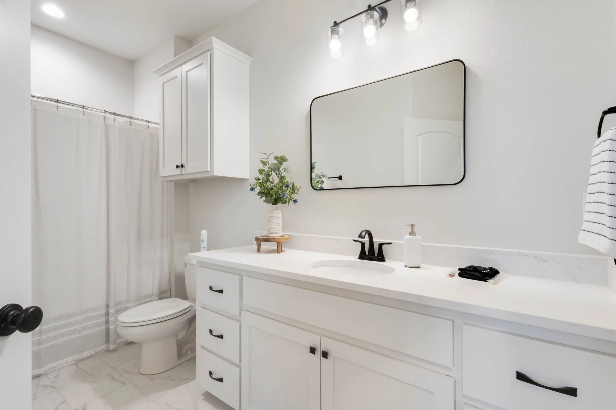 Modern white bathroom with a large mirror, a black faucet, a white countertop with a soap dispenser, a vase with flowers, and a cabinet. There is a toilet with a vertical window and a shower with a white curtain.