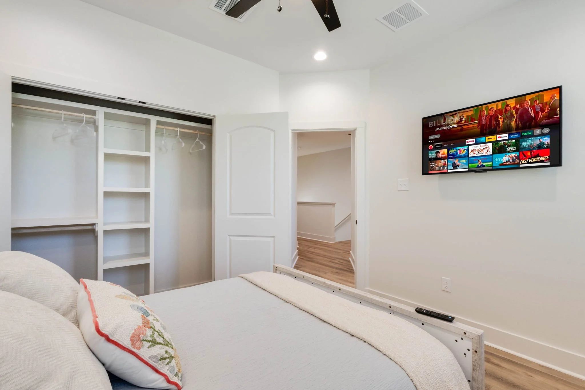 Bedroom with white walls, a mounted flat-screen TV displaying a streaming menu, a partially made bed with decorative pillows, an open closet with empty hangers, and a hallway visible through a doorway.