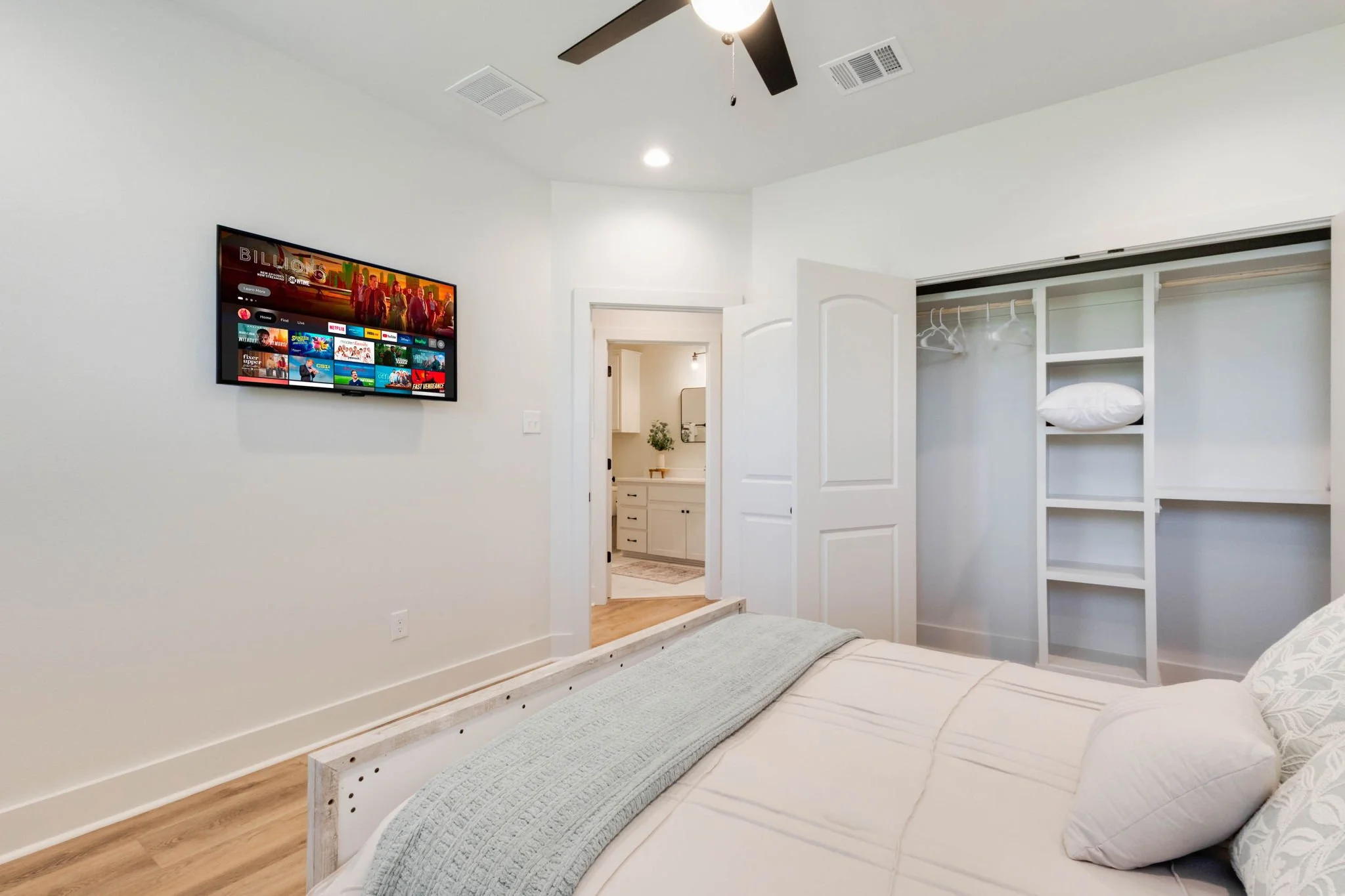 Bedroom with a mounted flat-screen TV, white walls, a doorway leading to a bathroom, an open closet with hanger rods and a pillow, a ceiling fan, and wood flooring.