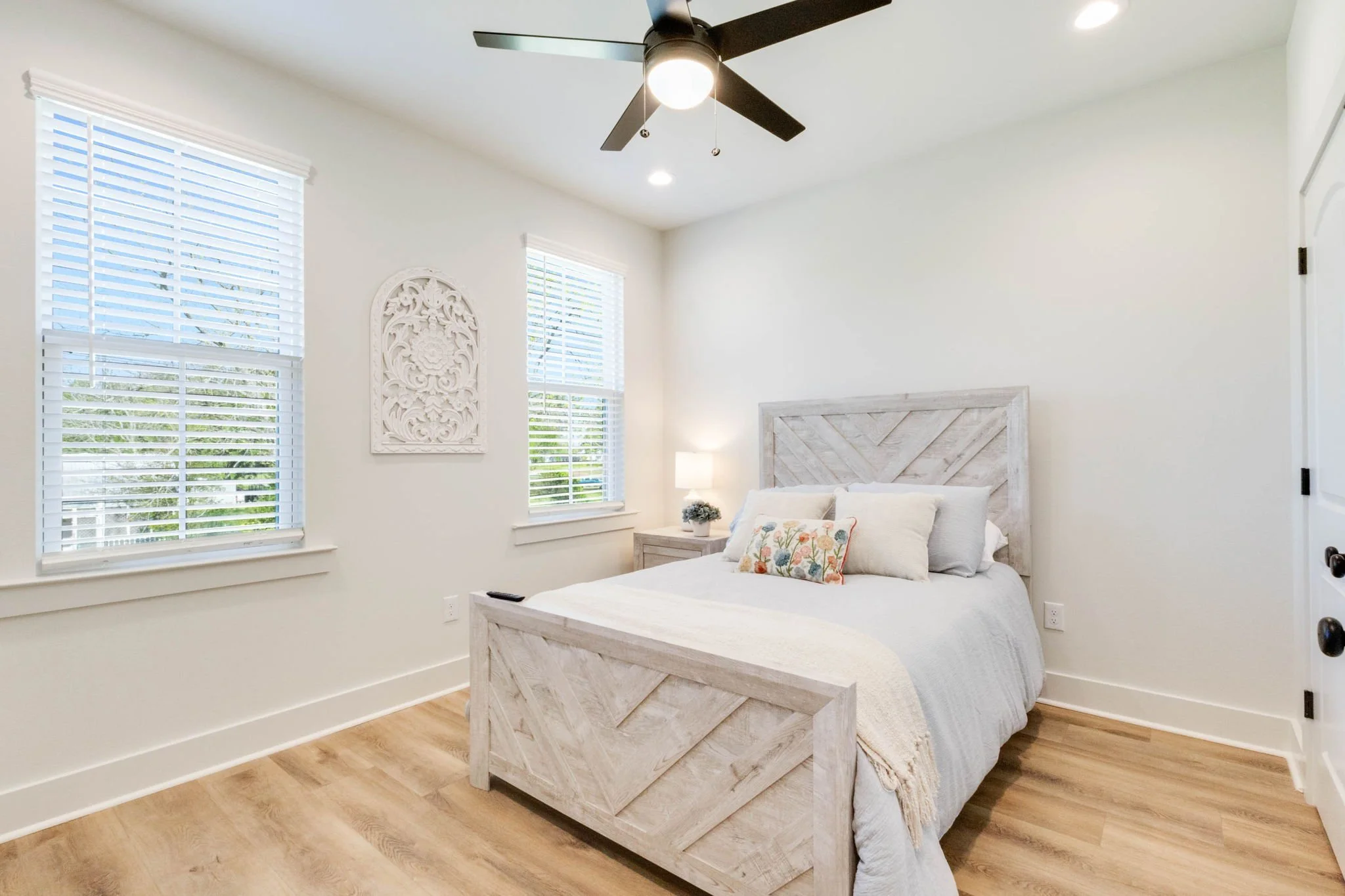 Bright bedroom with white walls, wooden flooring, a bed with a white headboard, pillows, a nightstand with a lamp, and windows with white blinds.