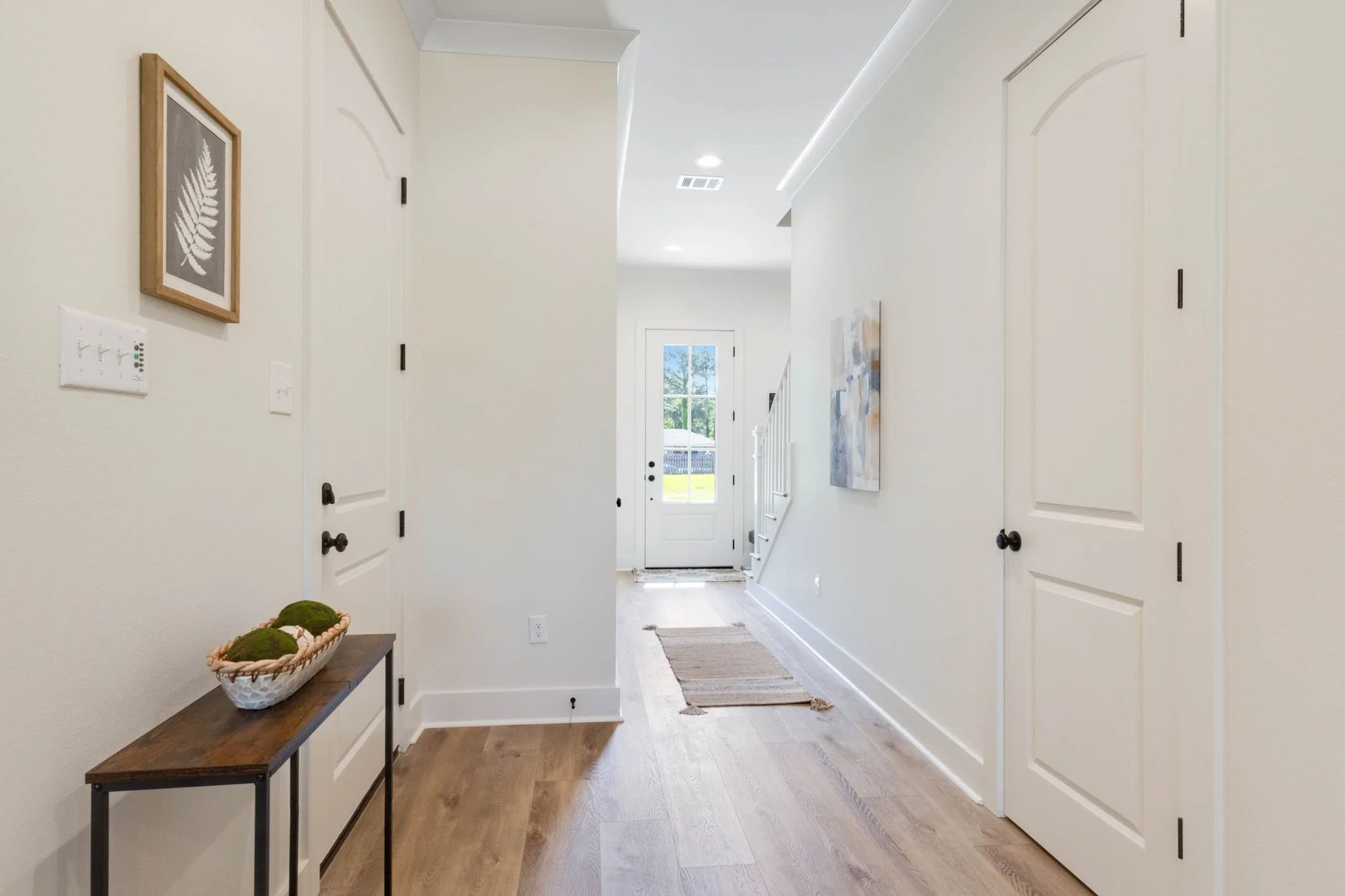 Bright hallway with light-colored walls, hardwood floors, artwork on the walls, a small table with decorative moss balls, and a door leading outside at the end of the hall.